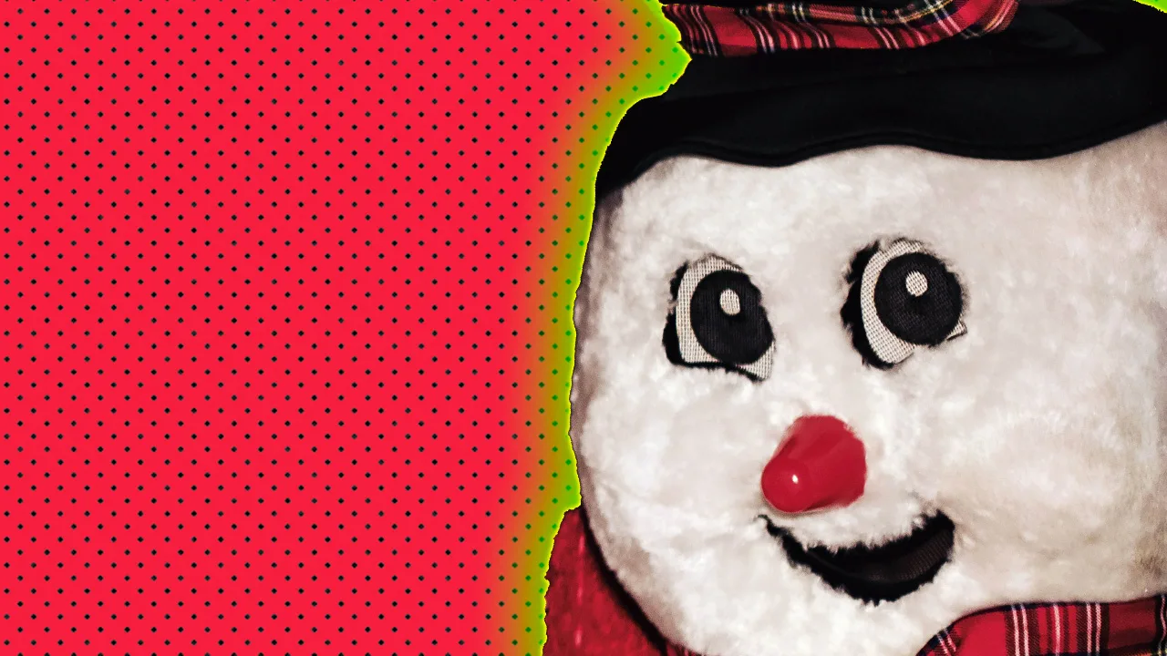 An image of a person dressed as a snowman with big eyes and a carrot for a nose. The snowman is wearing a tartan scarf and hat with a green halo against a red dotted background.