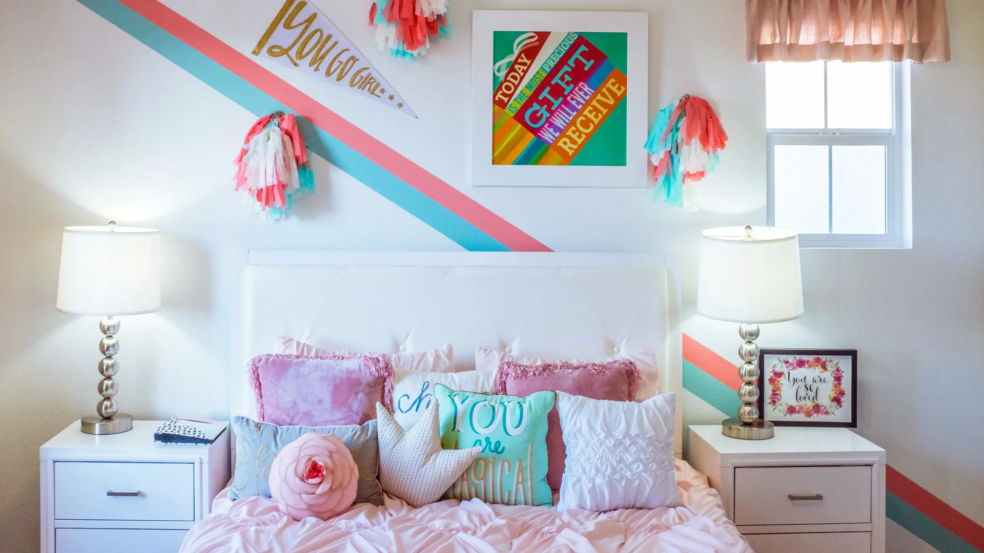 A photo of a colourful bedroom with pink green and white cushions on a pink bed with a colourful print in a frame on the wall with flags and pompoms. There are two bedside tables with silver and white lamps and a picture.