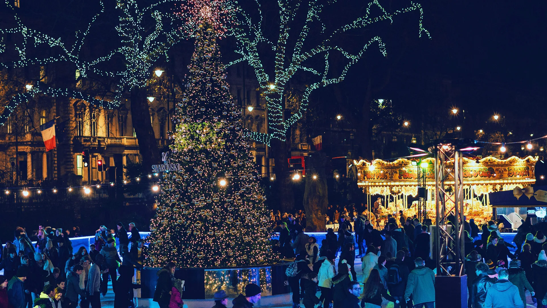 A photo of a Christmas tree all lit up at night in London with lots of people walking around it and a carousel next to it.