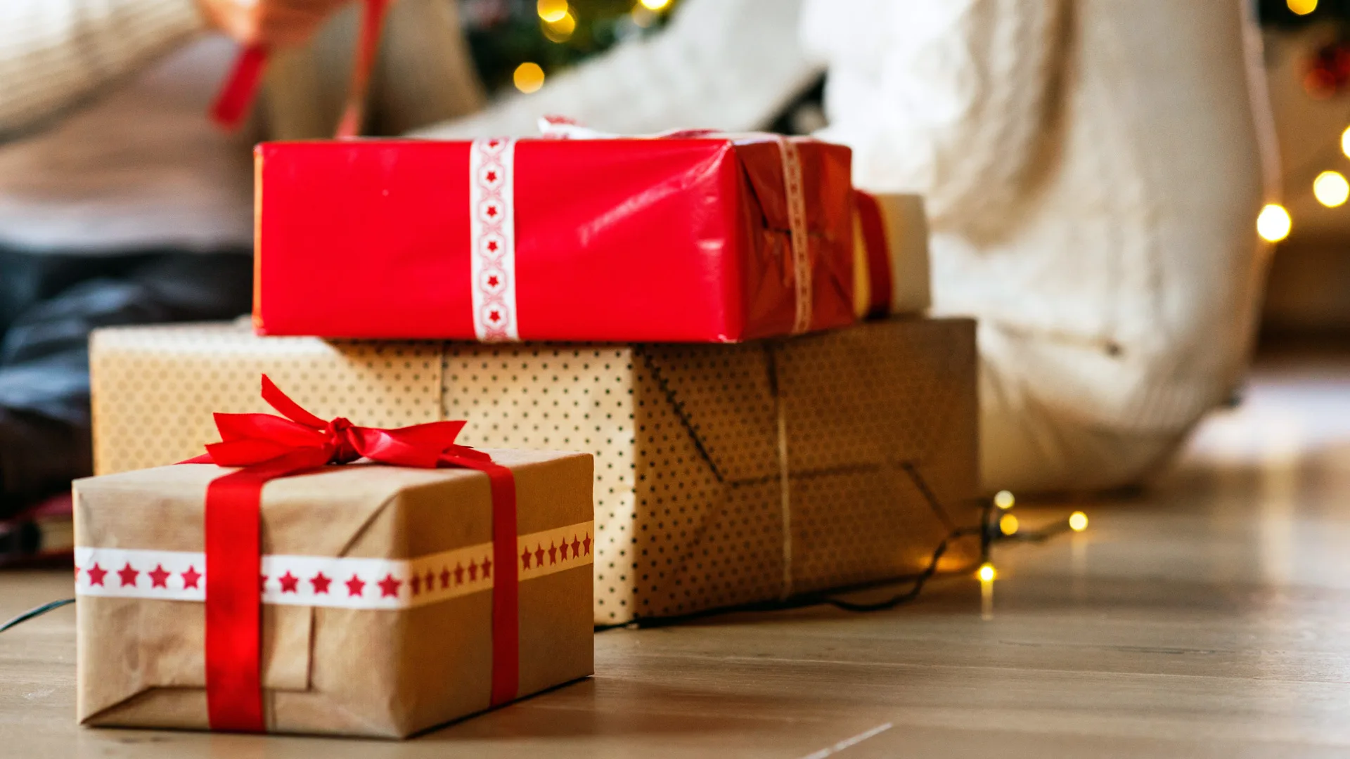 A photo of some giftwrapped red and beige presents on the floor with two people sitting nearby in jumpers near fairy lights.