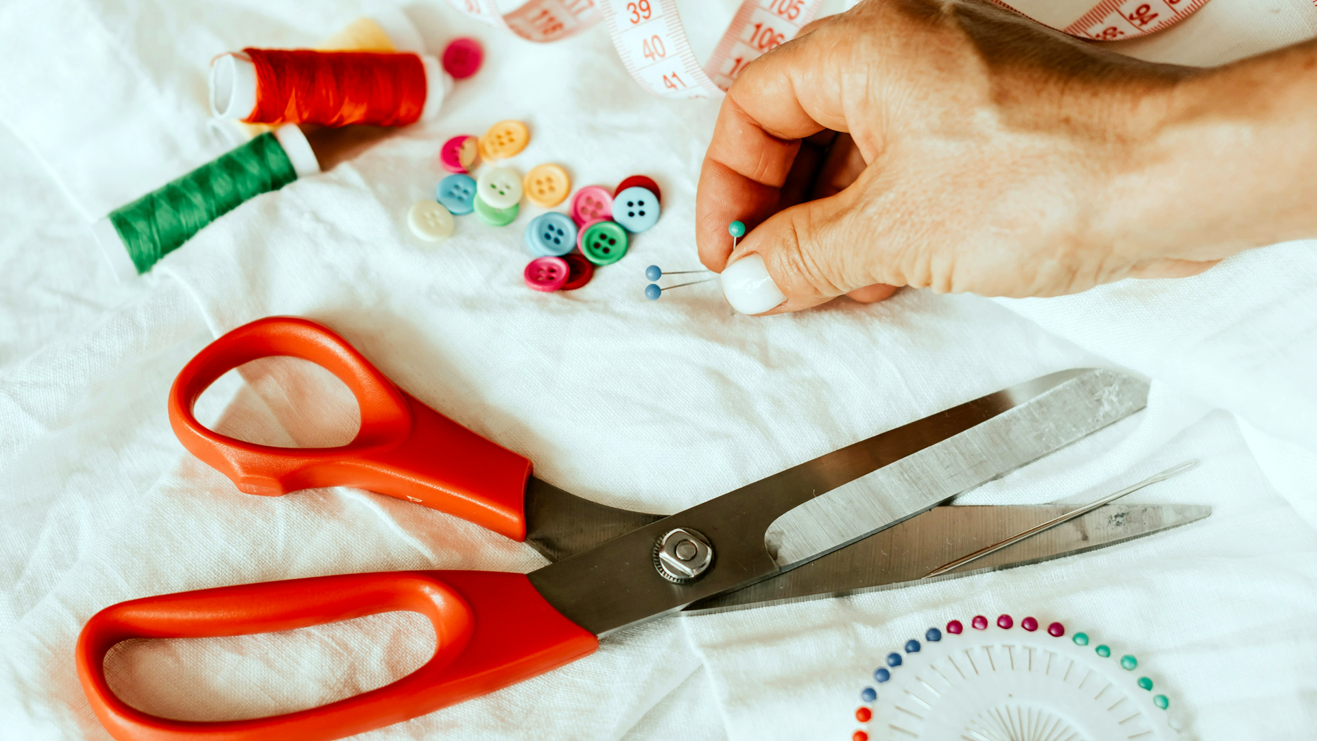 A photo of a hand sorting pins and buttons next to some orange handled scissors and more pins with red thread all lying on a white piece of fabric.