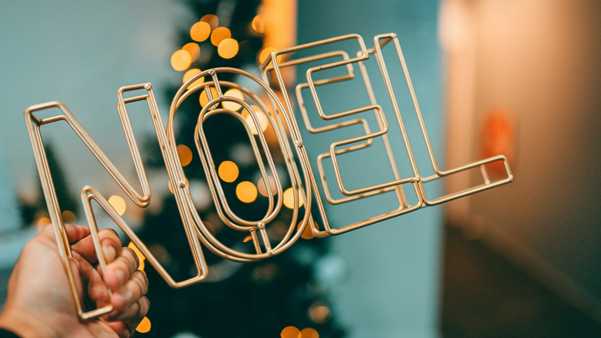 The words NOEL in gold wire being held by someone against a blue wall with an out of focus Christmas tree.