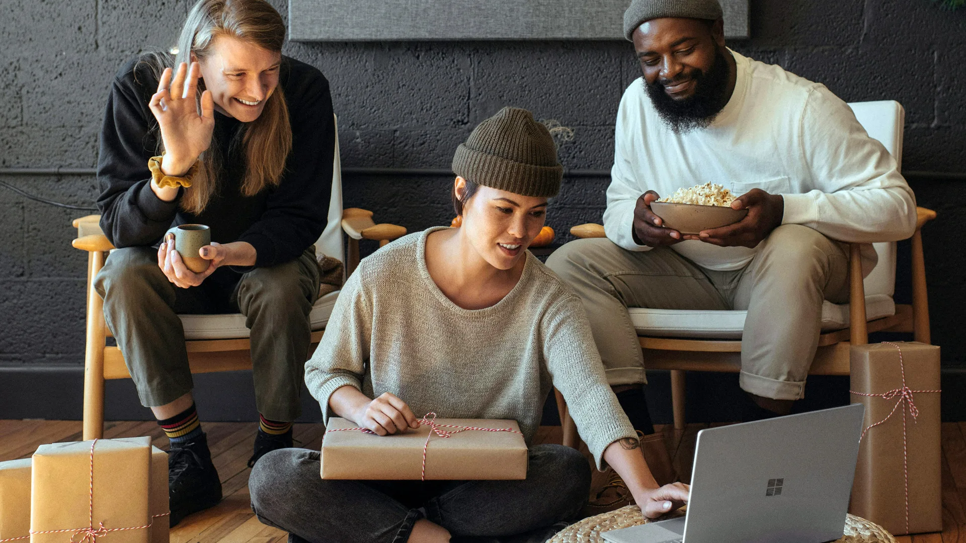 A photo of three people having a video call on a macbook. They are sat on chairs and smiling at the person on the call.