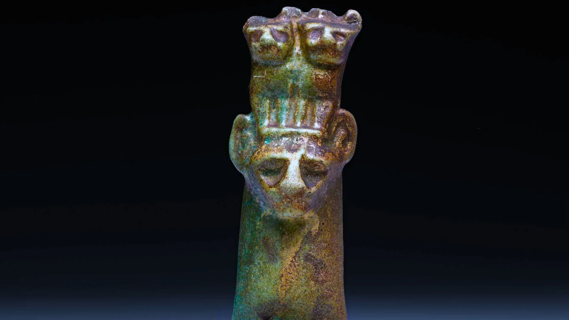 An image of an Ancient Egyptian amulet of the cat God Bastet in mixtures of greens, bronzes and dark brown - it is a cat head with two kittens sat on top against a black background.