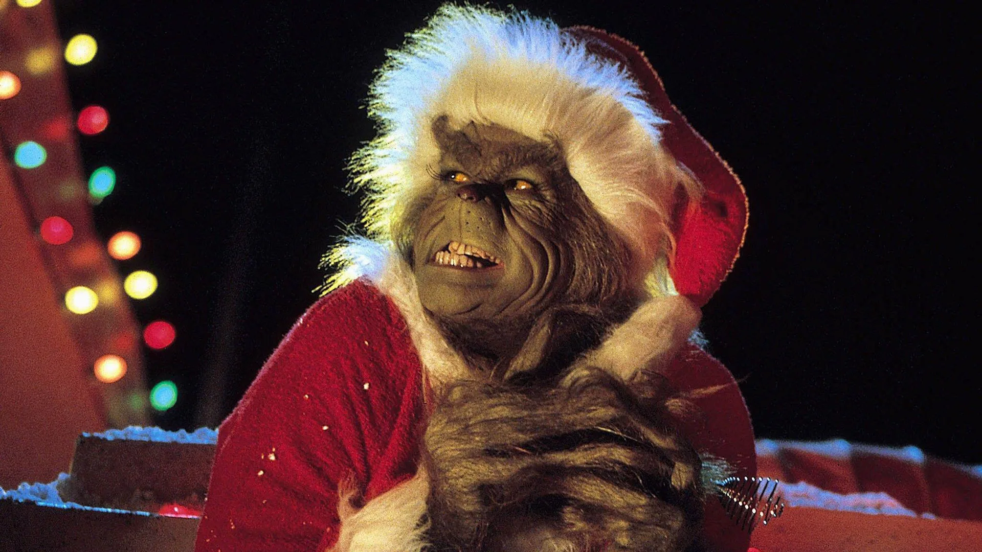 An image of The Grinch wearing a Santa outfit grimacing his face and holding his hands together against a dark background with Christmas lights.