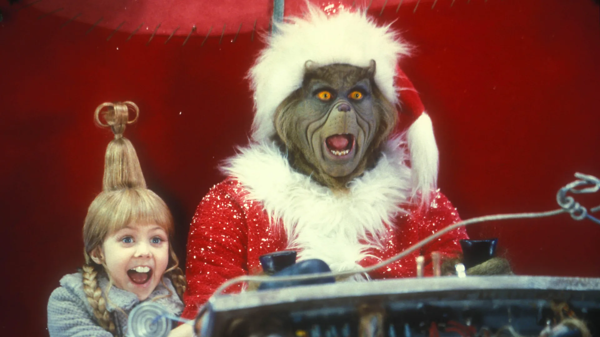 An image of The Grinch with Cindi Lou Who both screaming on a red sofa watching TV.