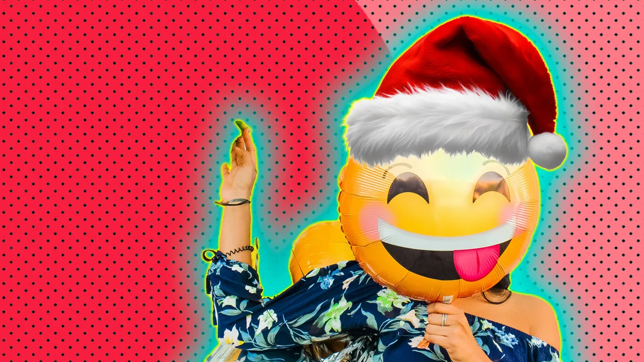 A photo of a woman holding an emoji balloon up covering her face. The balloon has a tongue sticking out expression and is wearing a Christmas hat with a blue and yellow halo against a red and pink dotted background.