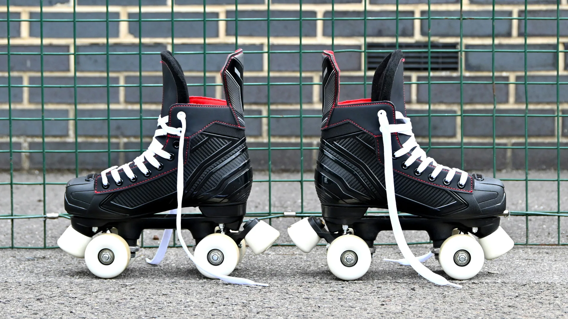 A photo of two rollerskates in black with white laces and wheels against a brick wall on a gravel ground.