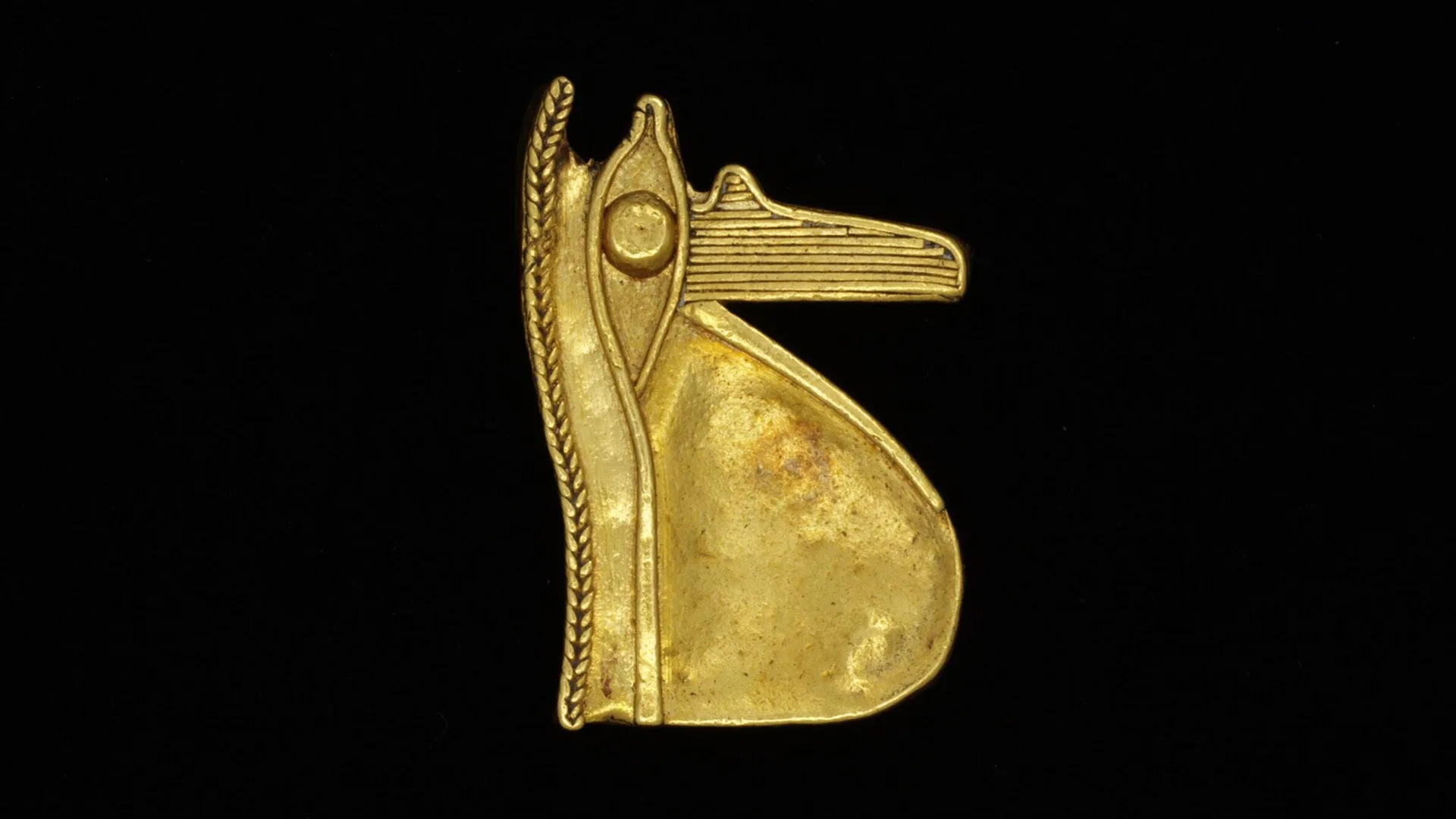 A photo of a gold amulet in the shape of Horus against a black background.