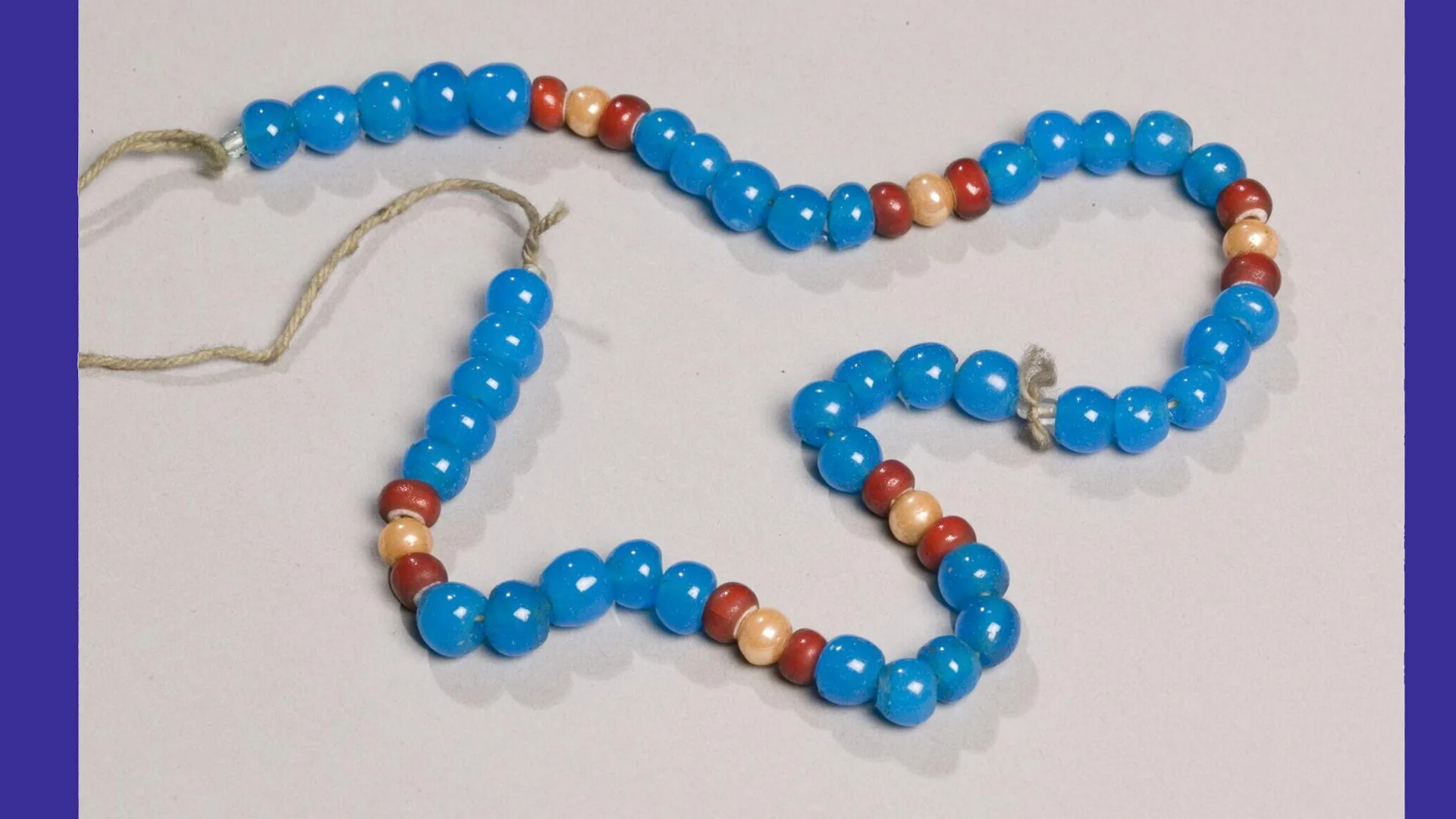 A photo of a glass beaded necklace in colours of blues, reds and gold against a grey background.