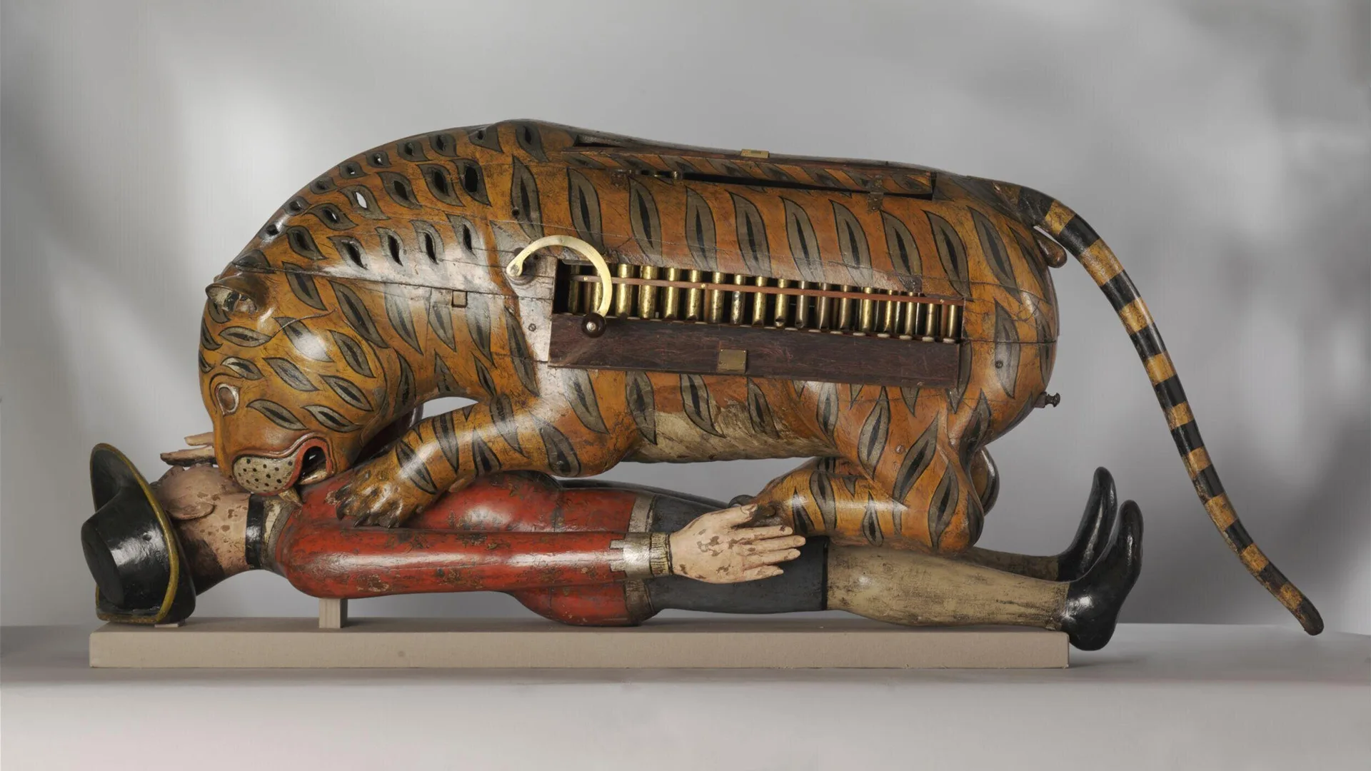 A photo of the object Tippoo's Tiger, an automaton tiger with an exposed in-built organ, on top of a British soldier in a red jacket whom the tiger is mauling. This is against a grey background.