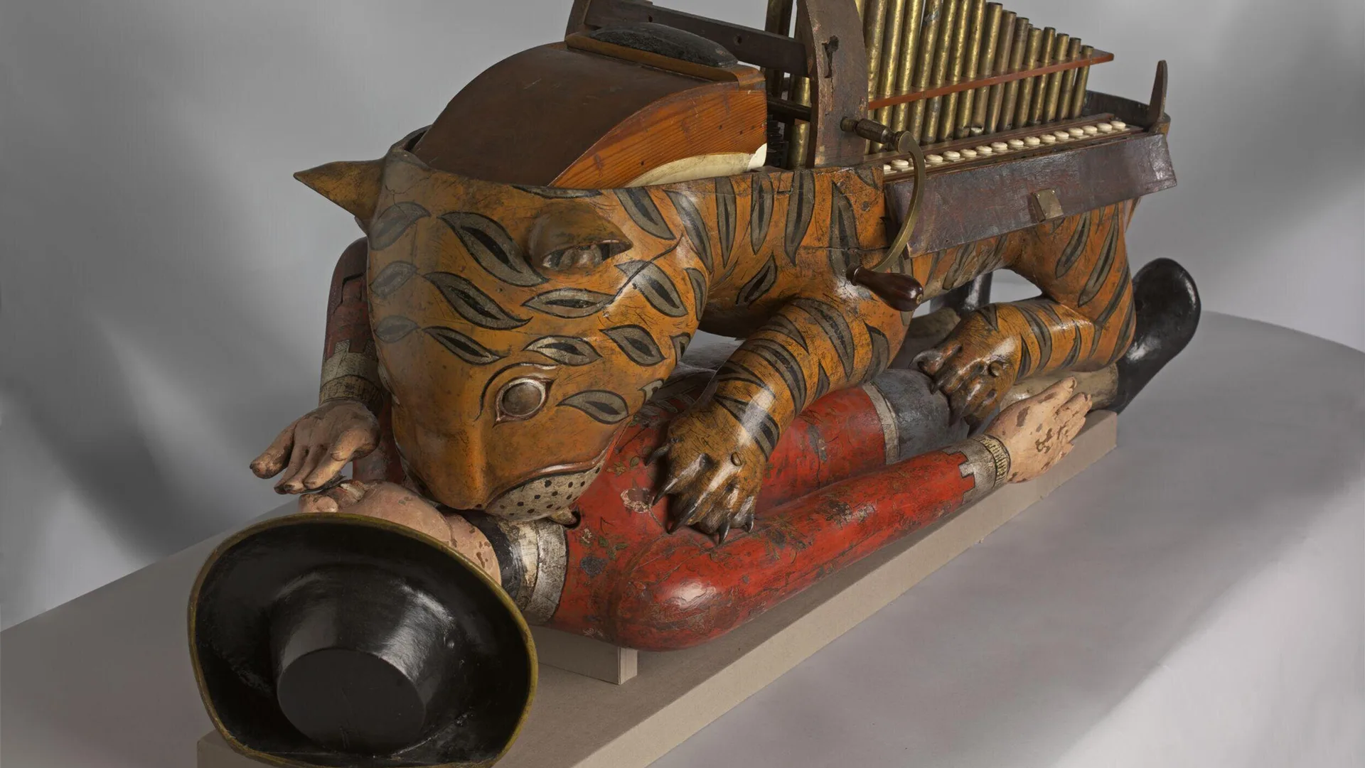 An image of the automaton Tippoo's Tiger showing a tiger with a built in organ exposed on it's side as it mauls a British soldier, lying on top of him as he screams. The tiger and soldier are on a grey floor.