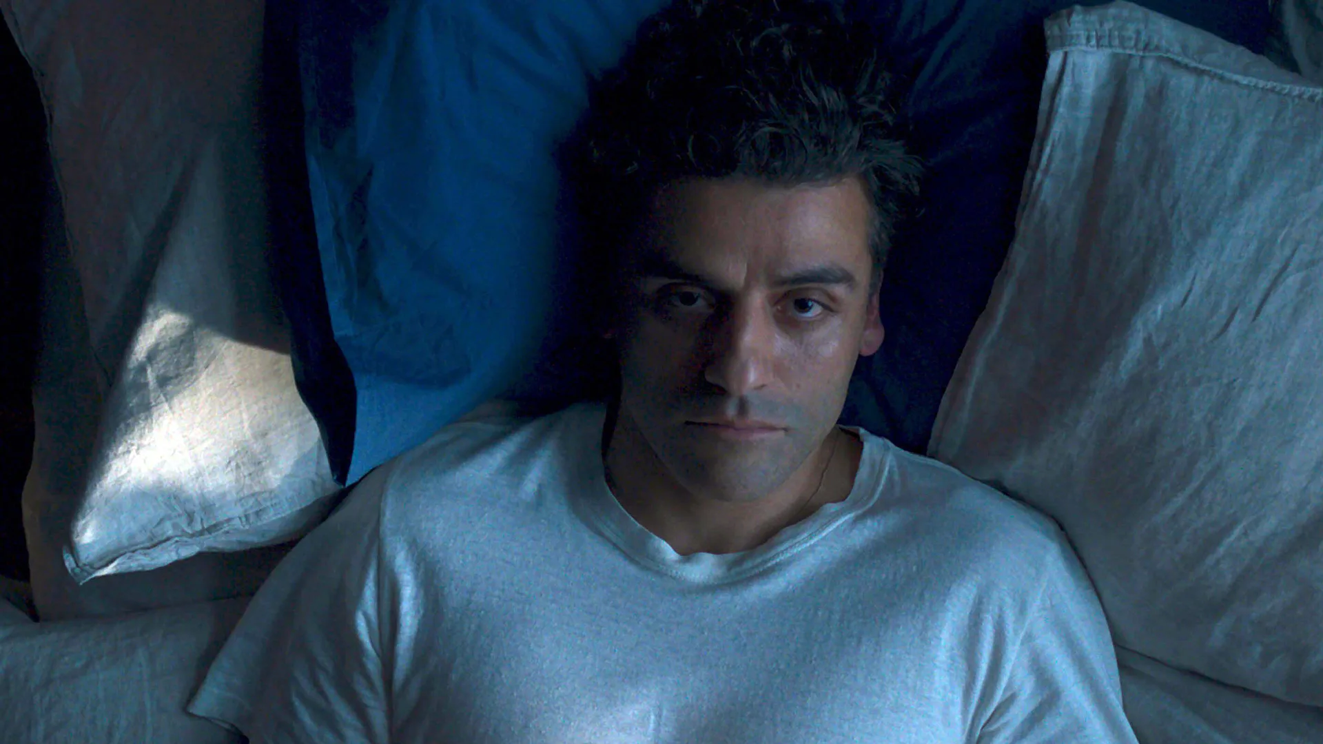 A photo of the actor Oscar Issac playing Steven Grant in Marvel's Moon Knight, lying on a bed looking at the ceiling He is wearing a white tshirt.