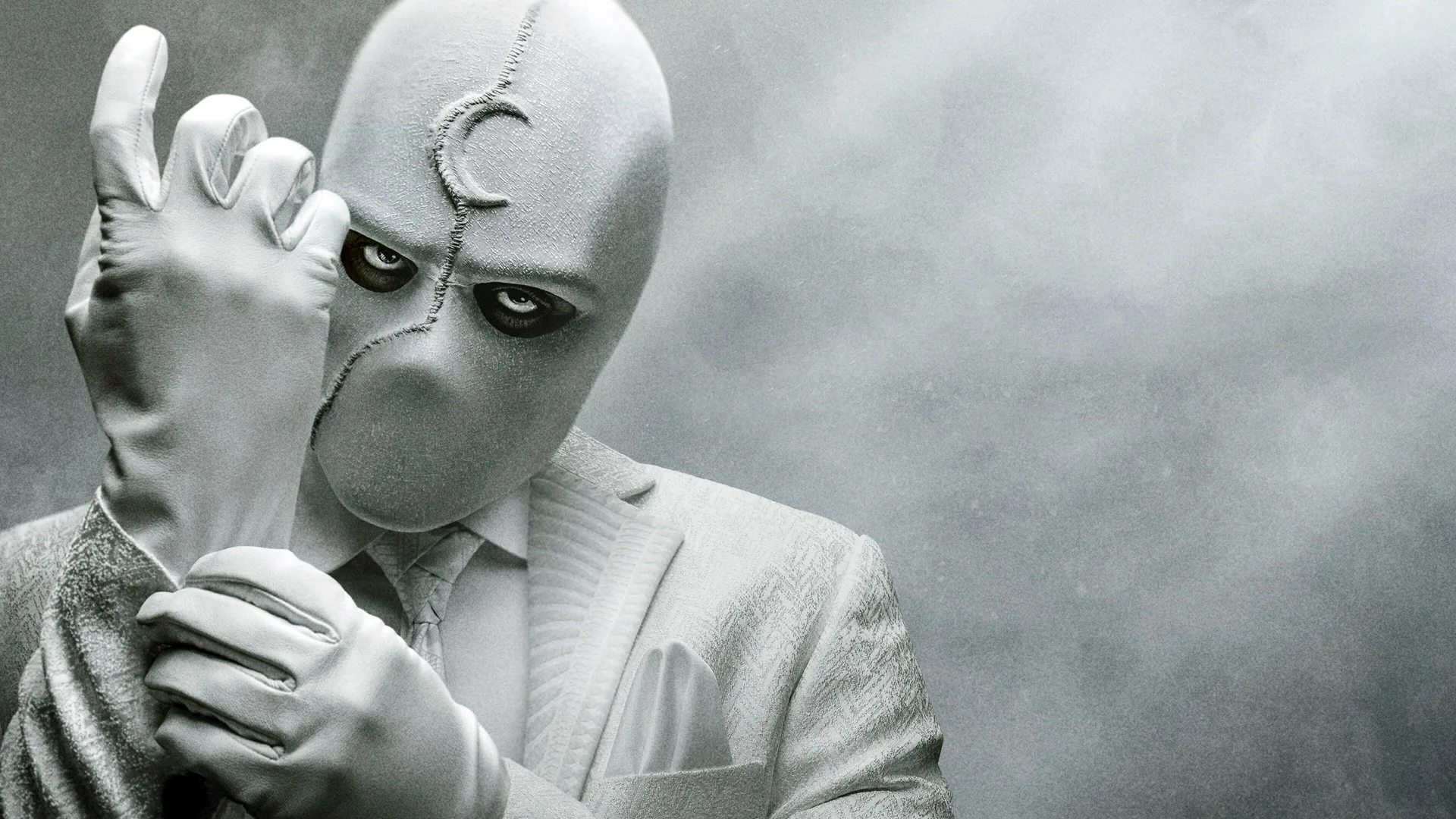 A photo of the Marvel superhero character Moon Knight, wearing a white jacket and gloves and a white head covering with a cresent moon on his forehead. He is holding up one hand and grabbing hold of the wrist with the other hand. He is against a grey cloudy background.