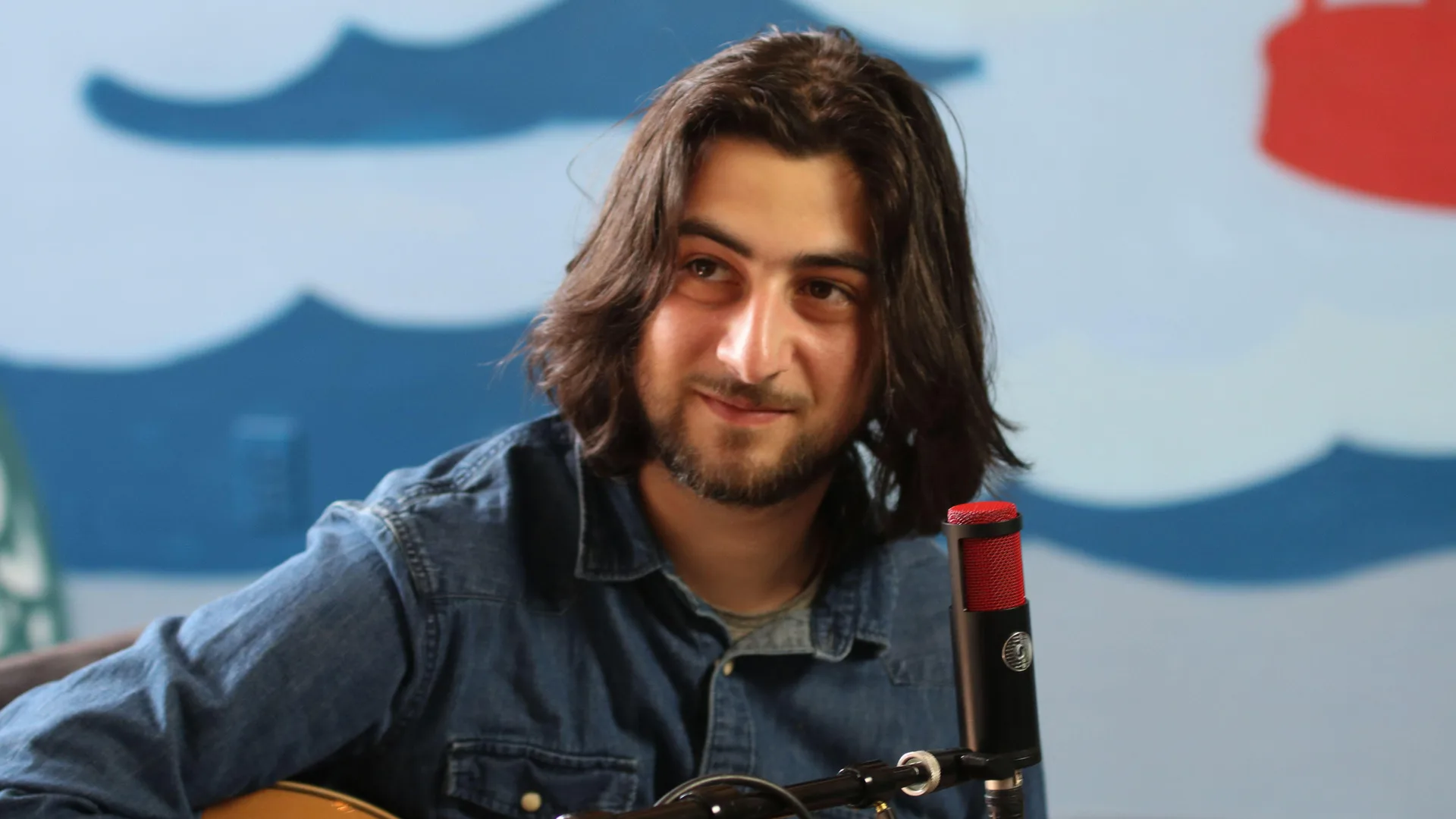 A photo of the singer Noah Kahan sat down with a guitar in arms wearing a denim blue shirt against a blue and white background.