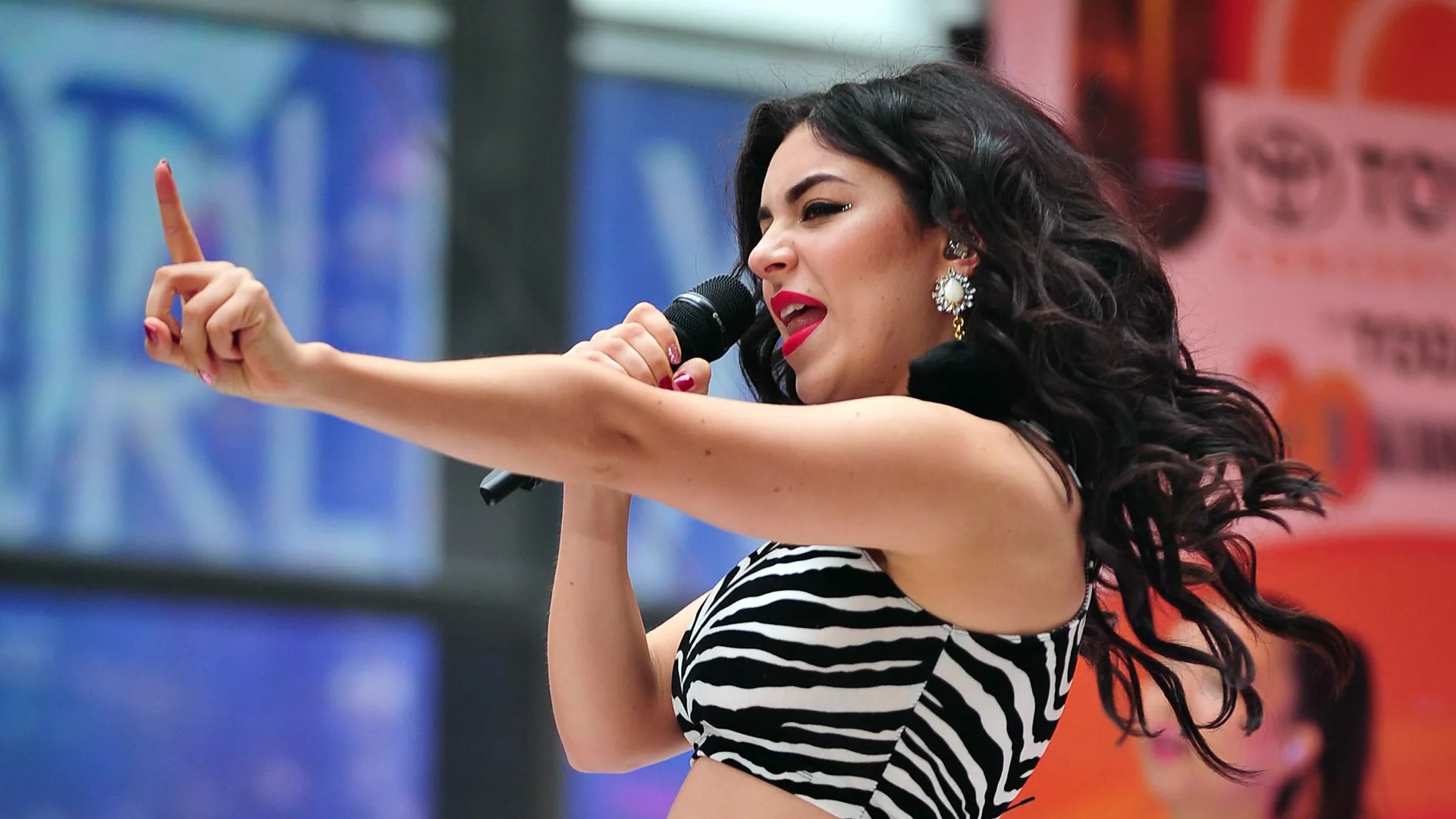 A photo of Charlie XCX singing on stage pointing her arm out waving her finger. She is wearing a zebra print top against a red and blue background of the stage.