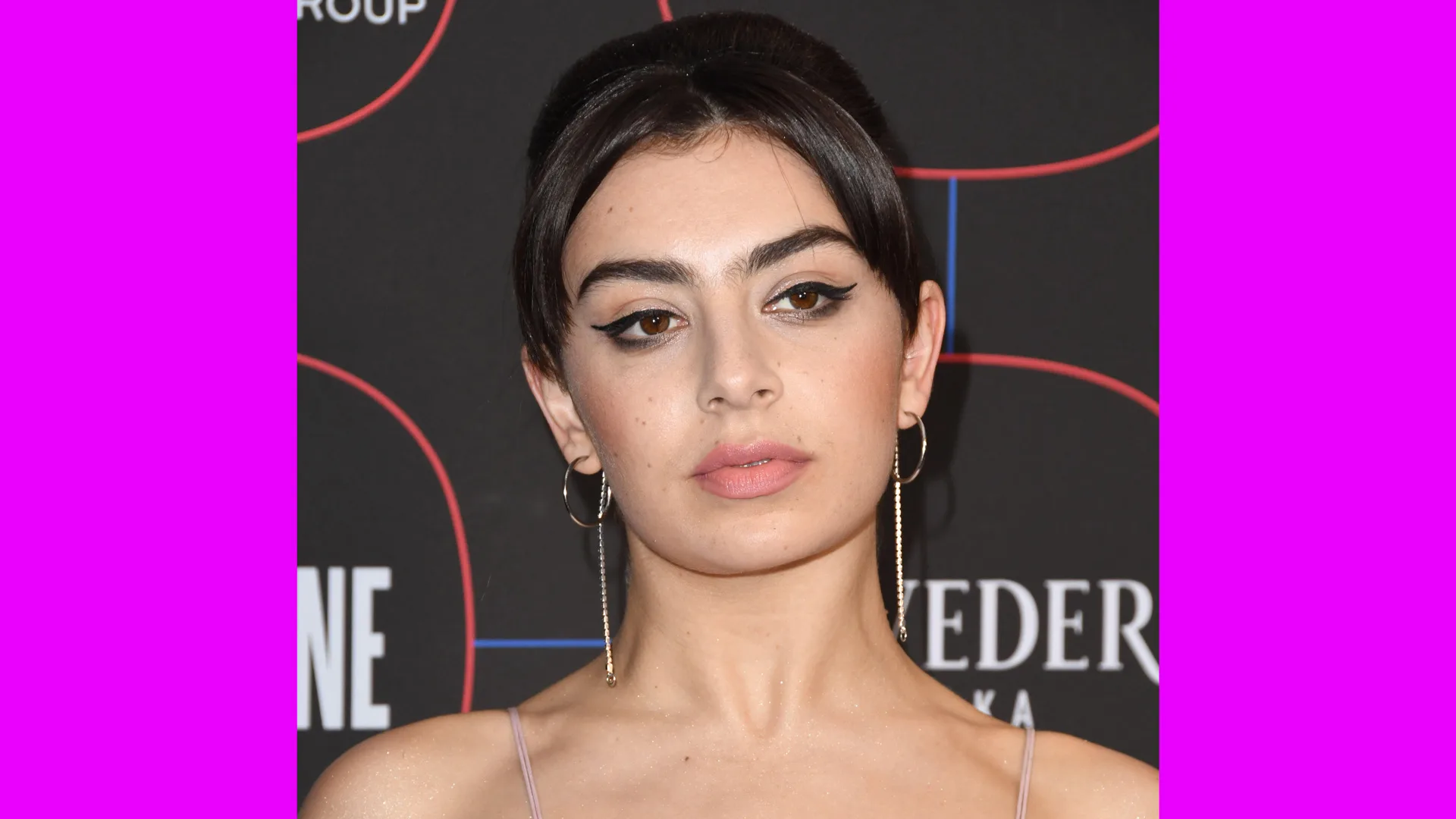 A photo of Charlie XCX looking off to the distance wearing a dangly pair of silver earrings and her dark hair centre parted and up against a black advertising billboard with orange stripes, bordered with pink either side.