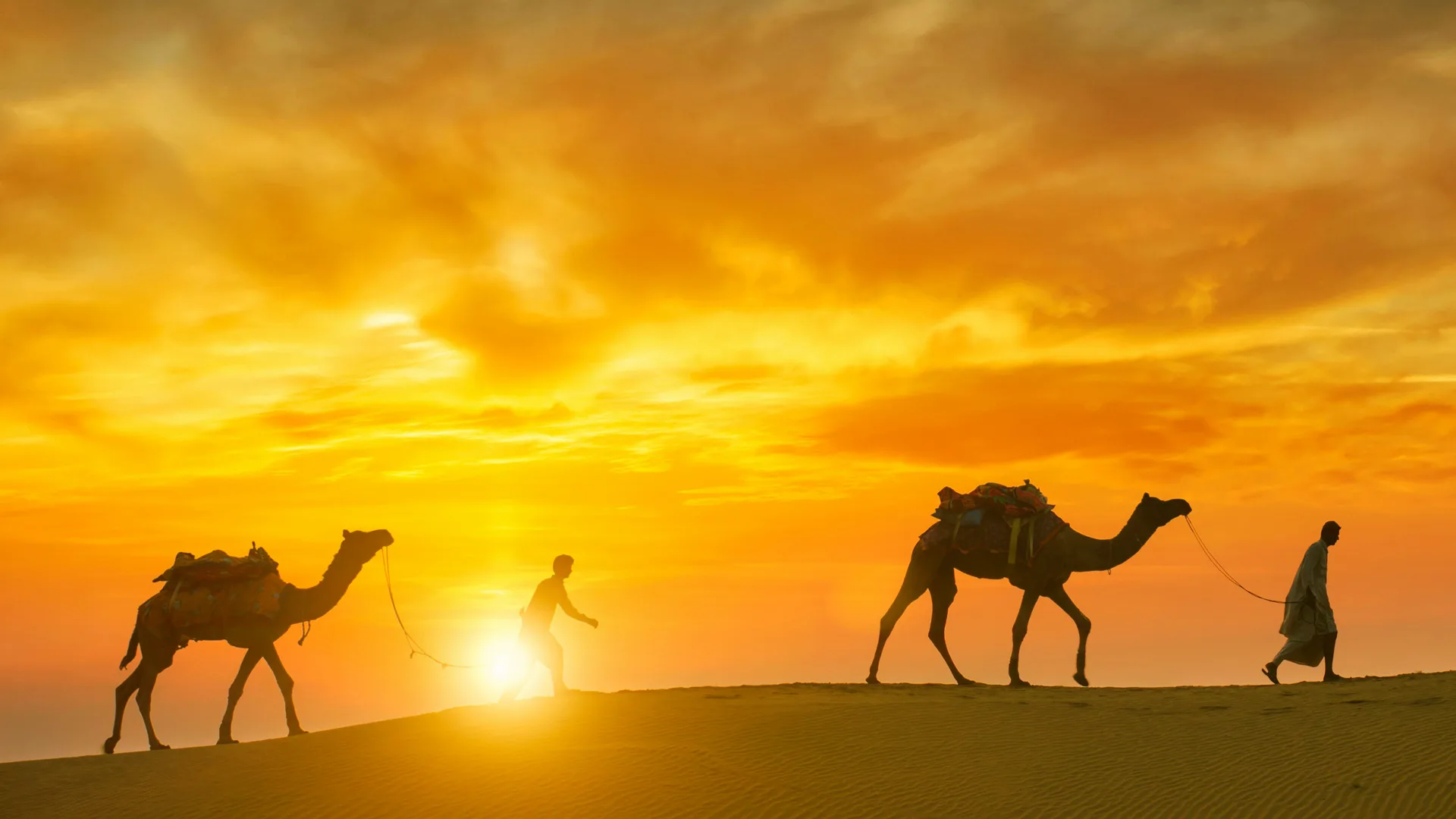 A photo of a sunset with bright yellow sky and the silhouette of two camels and their herders walking along the desert horizon line.