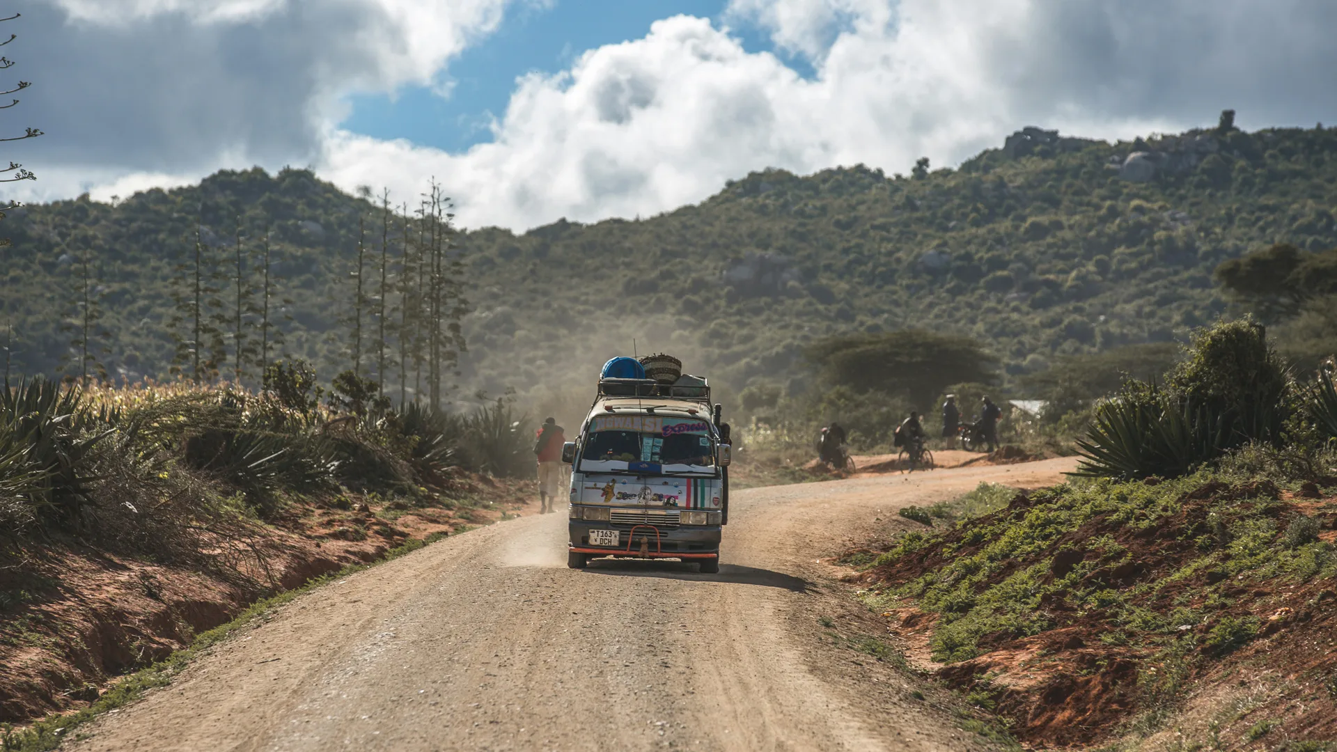 A dirt road with a bus driving in the distance with green hills and blue sky in the background.