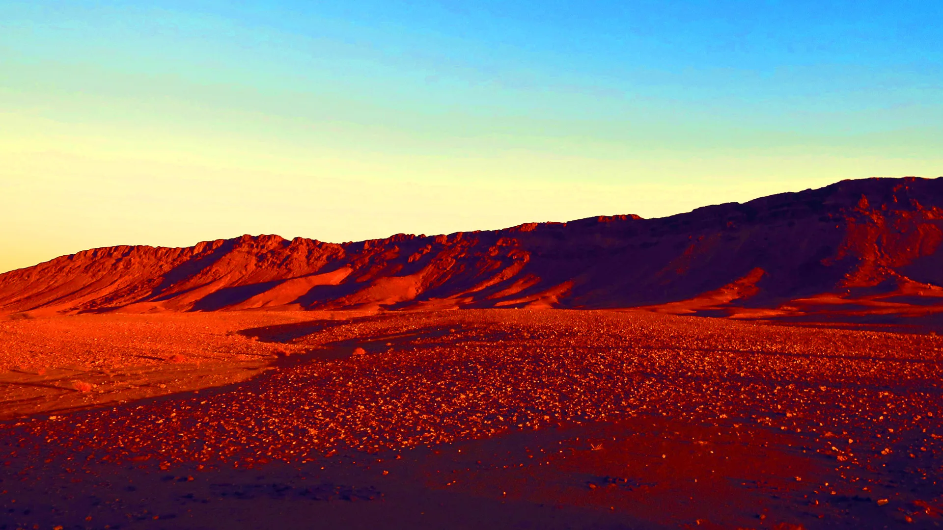 A photo of the desert in different tones and shades of reds with a blue and yellow sky.