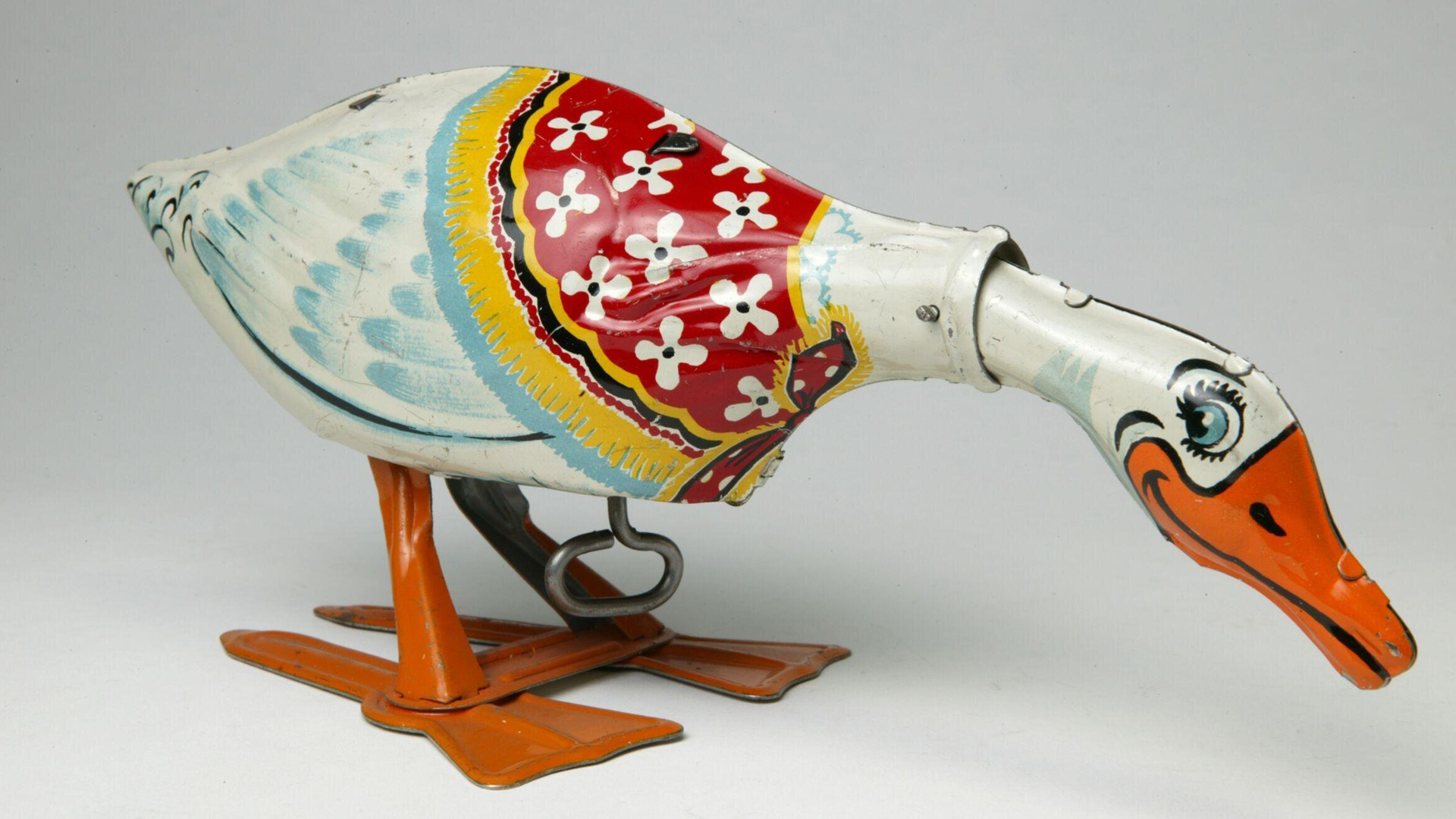 A photo of a clockwork toy goose with a smiling beak and cartoon eyes bending over wearing a little red and yellow shawl with white flowers.