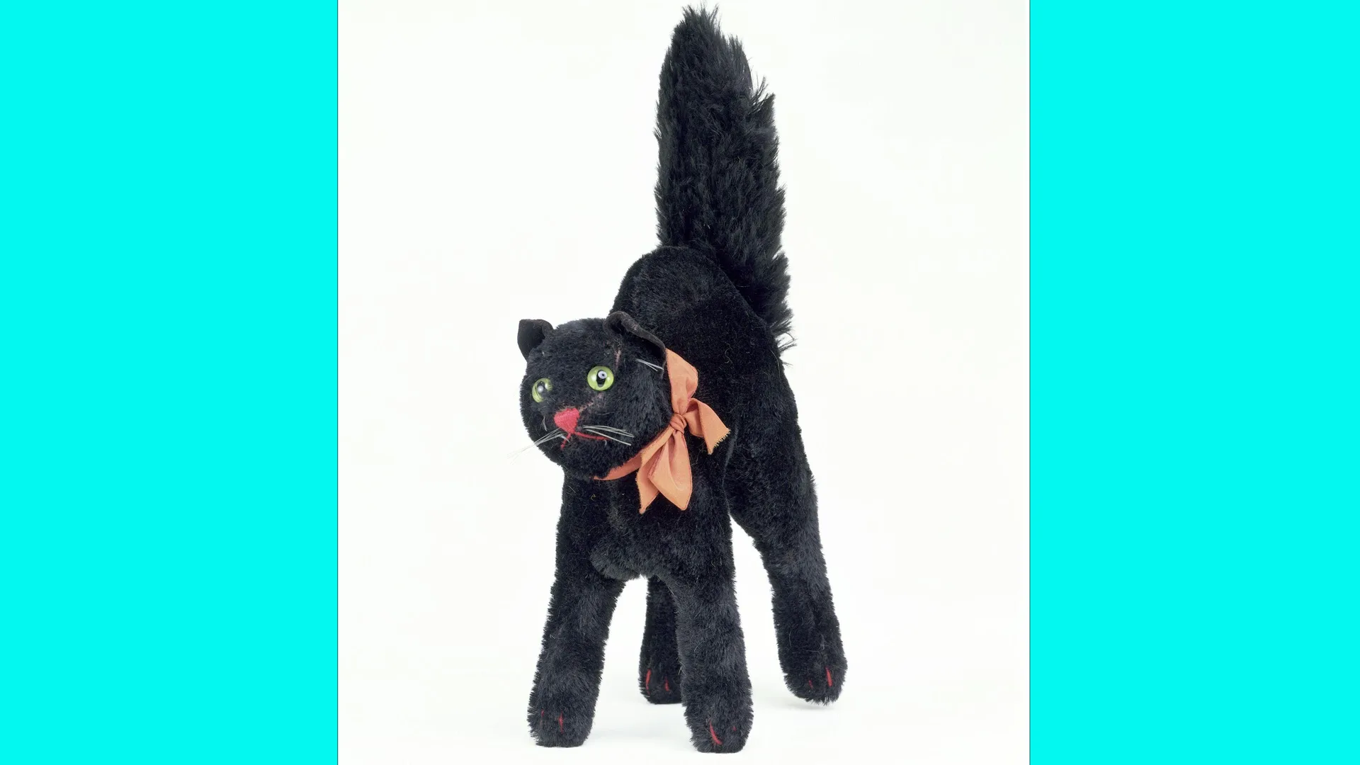 A photo of a black toy cat with it's tail stretched upwards, wearing a pink ribbon around its neck with bright green eyes and a pink nose, standing on a white surface with blue borders.