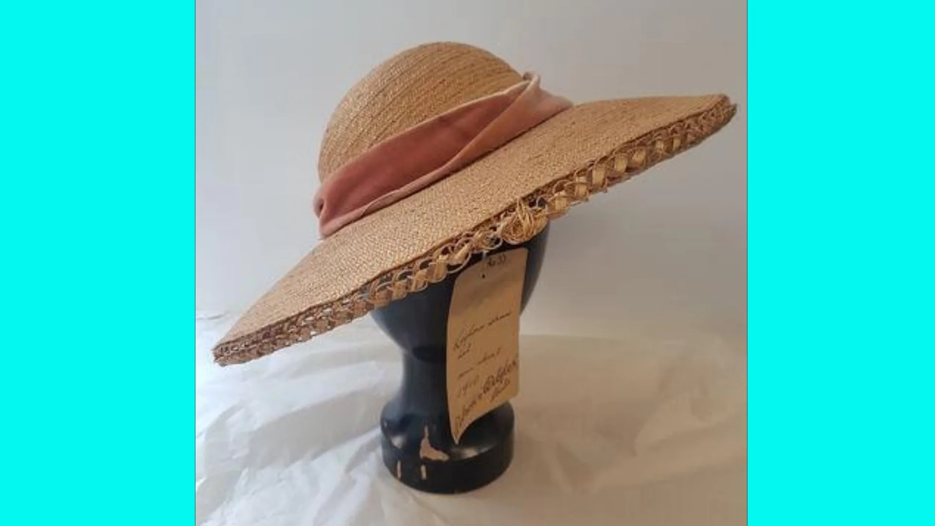 A photo of a wide brimmed straw hat with a pink velvet ribbon around the base and a price tag hanging from the hat which is mounted on a black head mannequin, sat on a white cloth surface with blue borders.