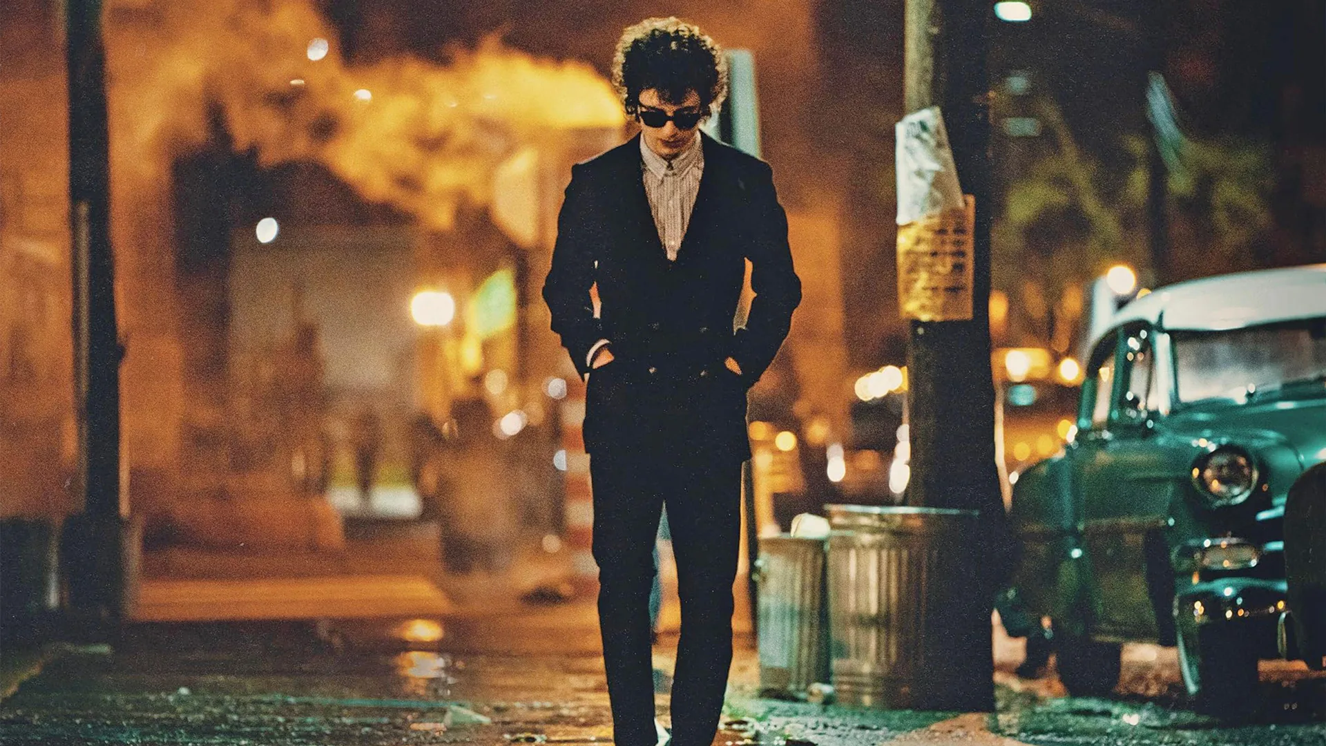 A still from the movie A Complete Unknown showing the actor playing Bob Dylan walking down a street with gold and blue lighting.