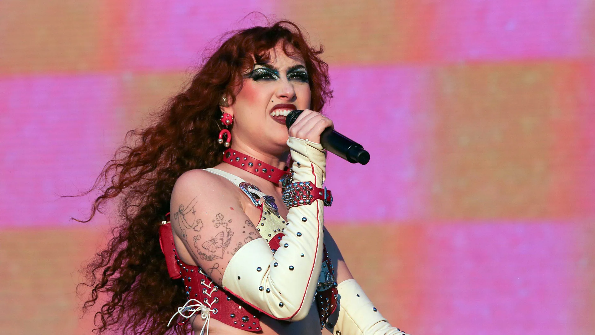 A photo of Chappell Roan performing on stage singing into a mic, wearing white leather studded gloves and a red corset top with pink and yellow squares behind her.