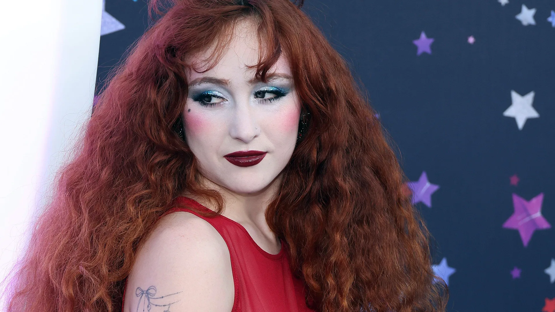 A photo of Chappell Roan smiling to the side wearing dark lipstick and silver grey eyeshadow with a red top showing her shoulder tattoos. Behind her is a dark blue background with white and pink stars.