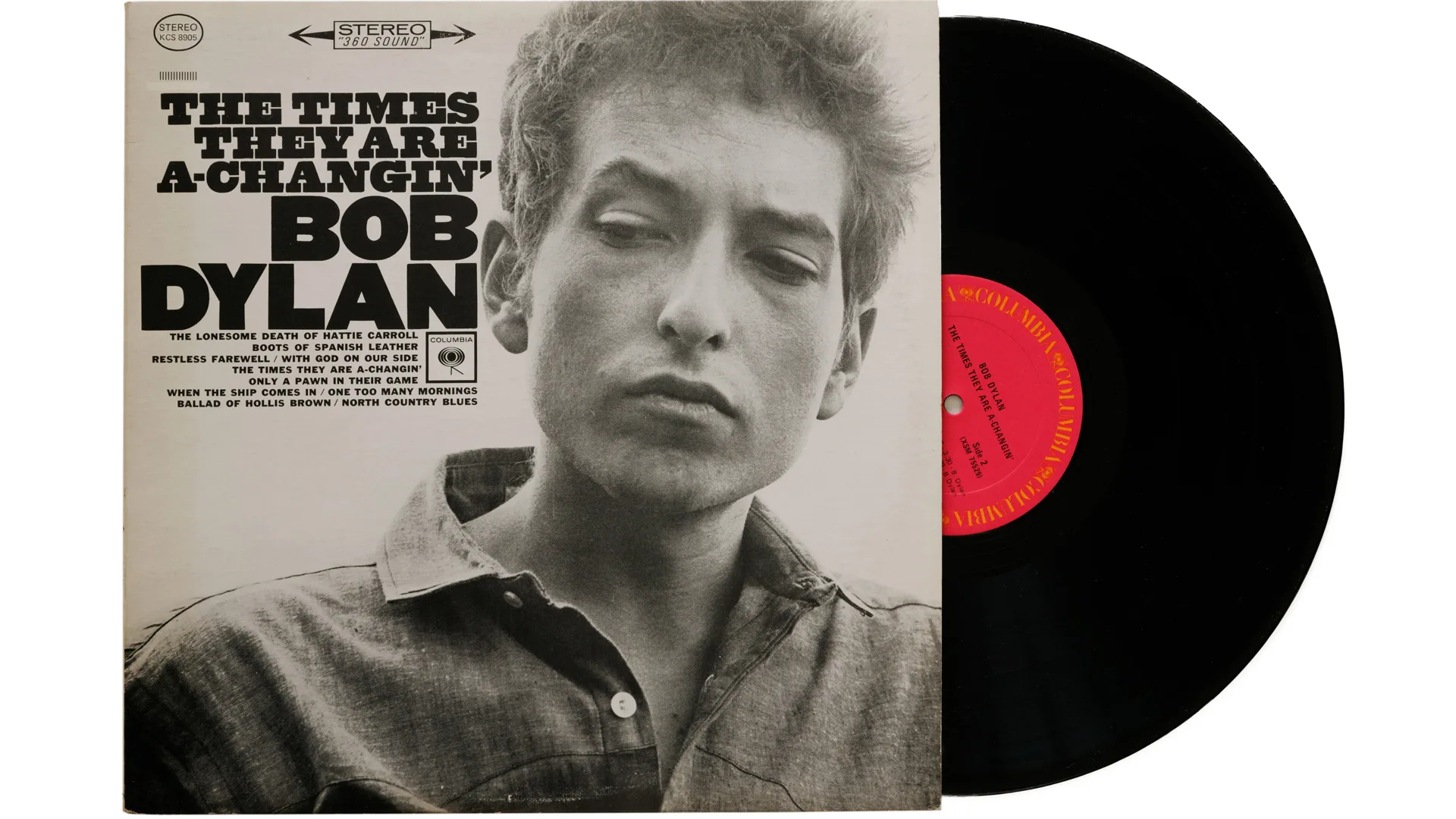 A photo of the Bob Dylan album "The Times They Are A-Changin'" on vinyl showing Bob Dylan in black and white look to the side wearing a shirt and the black record poking out from the side with a red label - all against a white background.