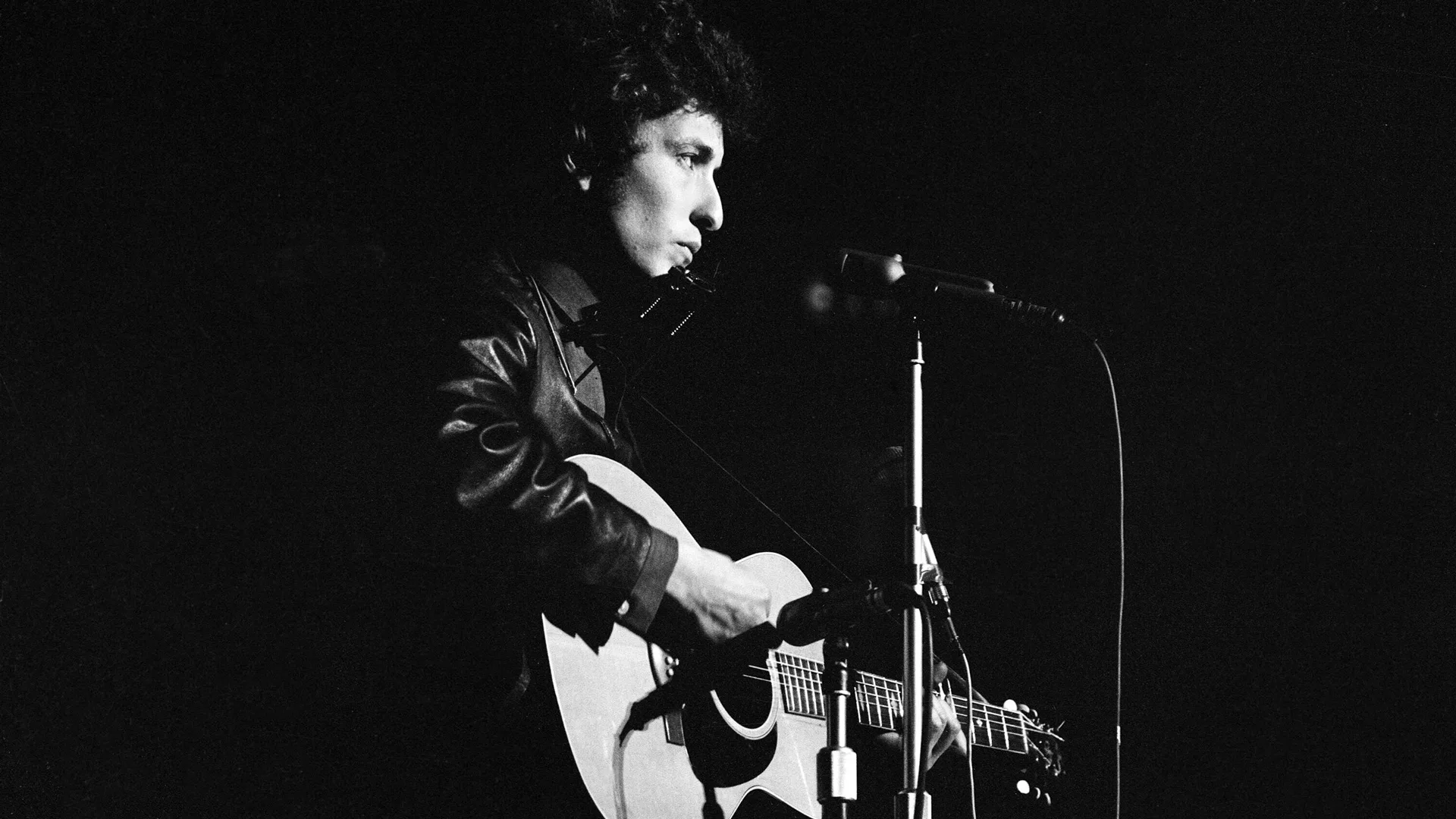 A black and white photo of Bob Dylan stood on stage at the Royal Albert Hall performing with his guitar and mic.