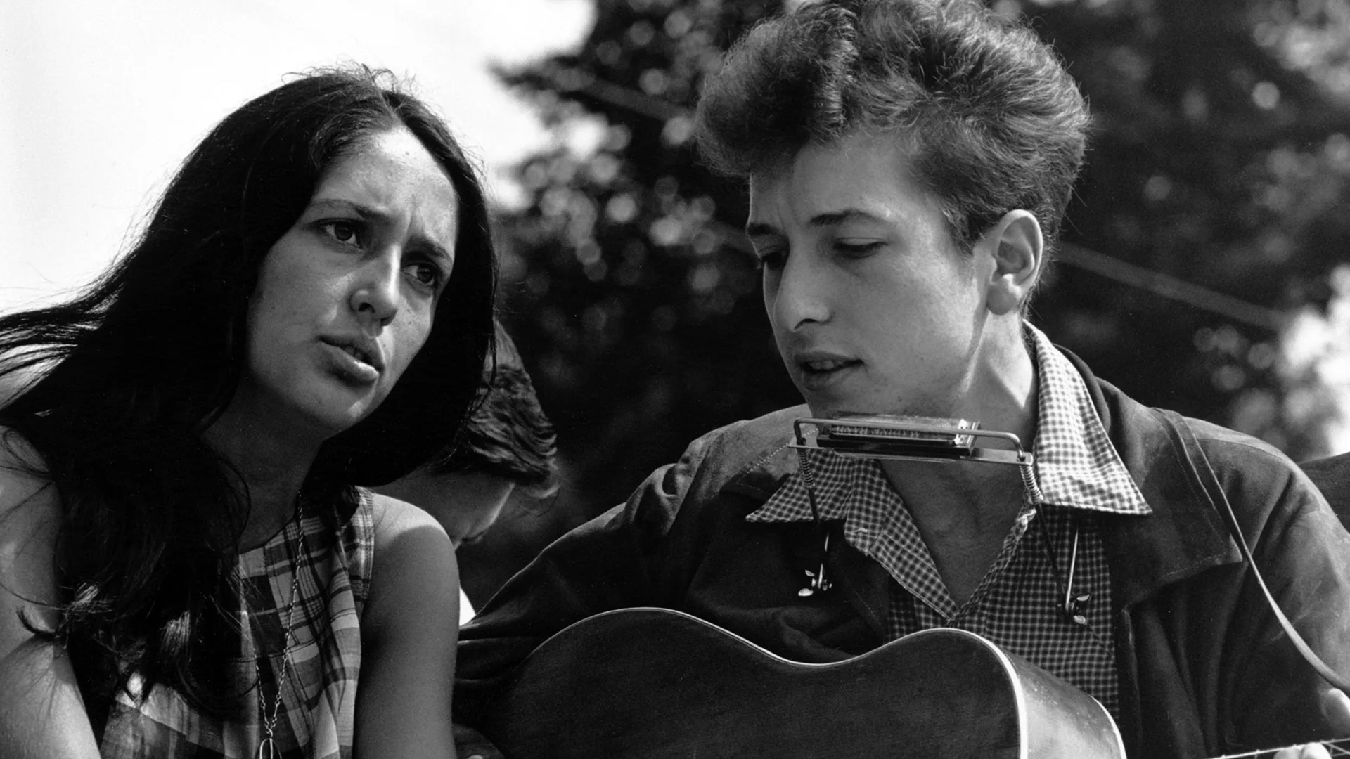 A black and white photo of Joan Baez and Bob Dylan at the March on Washington in 1963. He is singing into a mic with a guitar and she is looking out at the crowd. Behind them are trees.