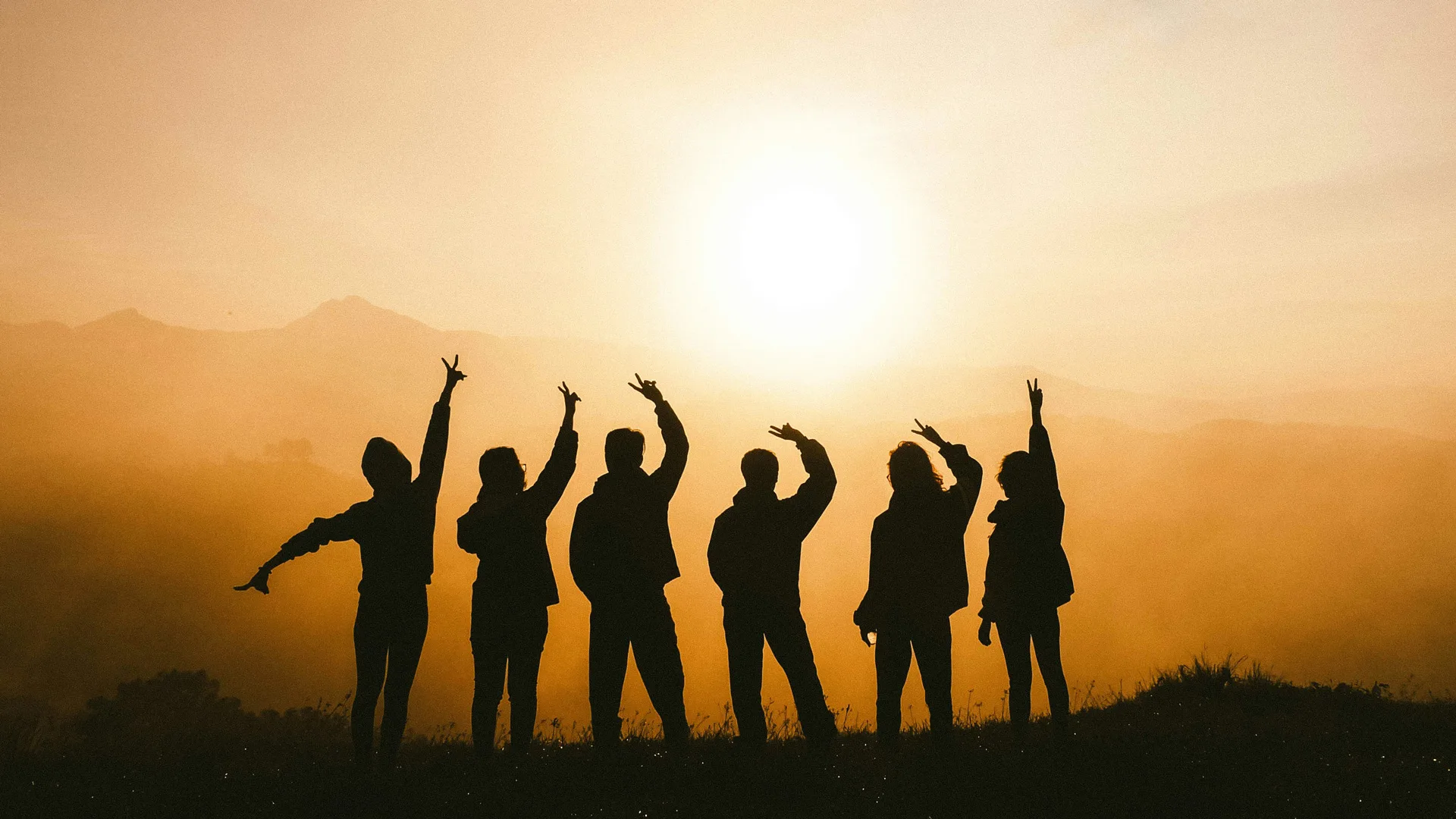 A photograph of a group of people stood on a hillside as the sun sets orange above them. They are silhouetted and holding their arms in the air.