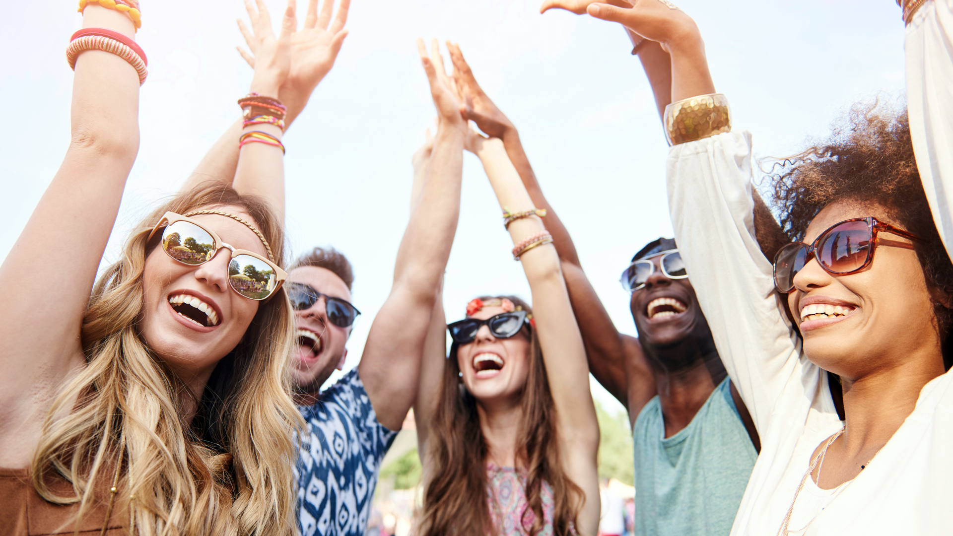 A photo of a group of young people wearing sunglasses with their hands raised in the air laughing. The sky is brightly lit behind them.