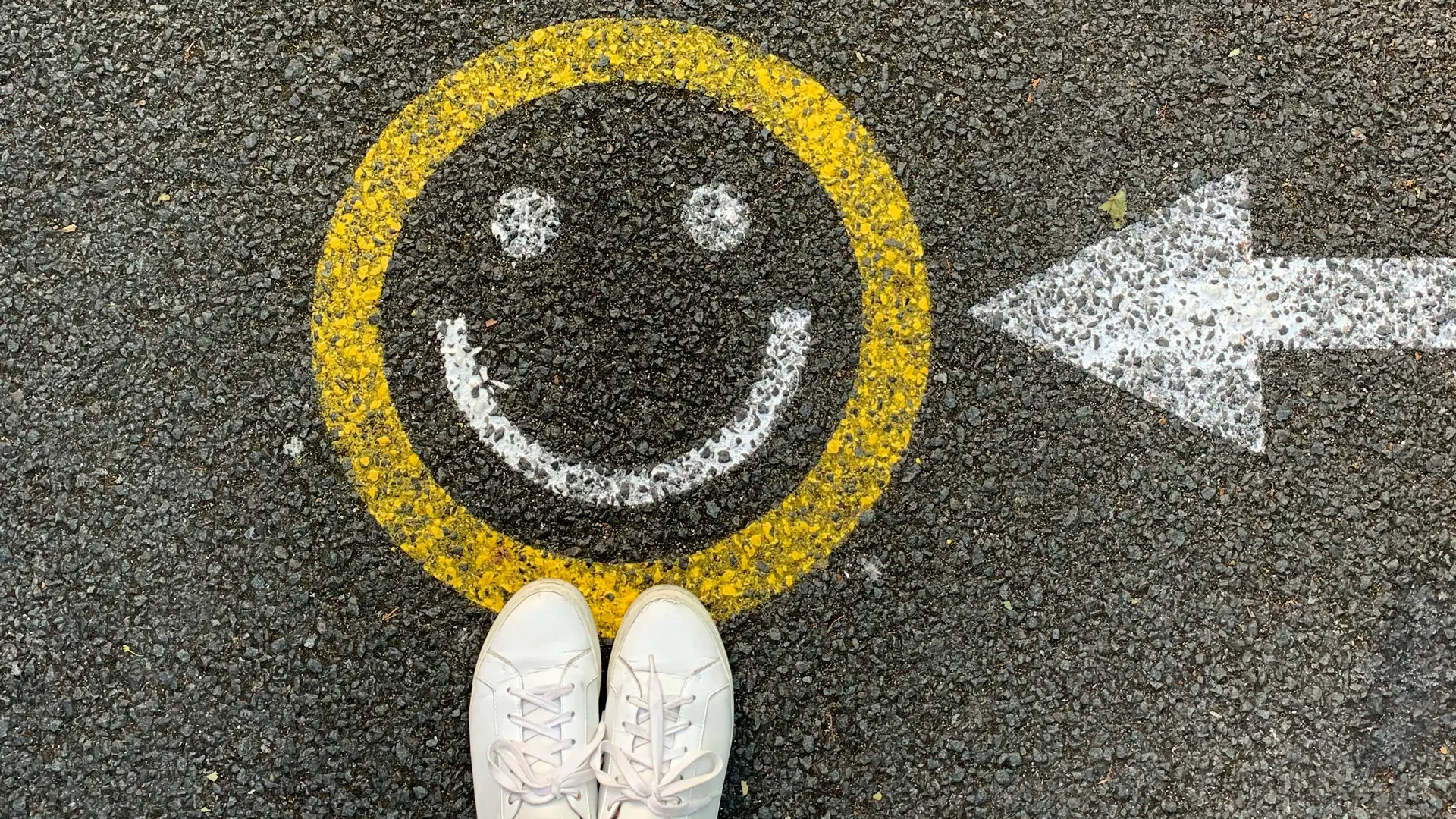 A photo of a ground with a painted smiley face with white eyes and mouth and a yellow circle and a pair of white trainers together standing on it.