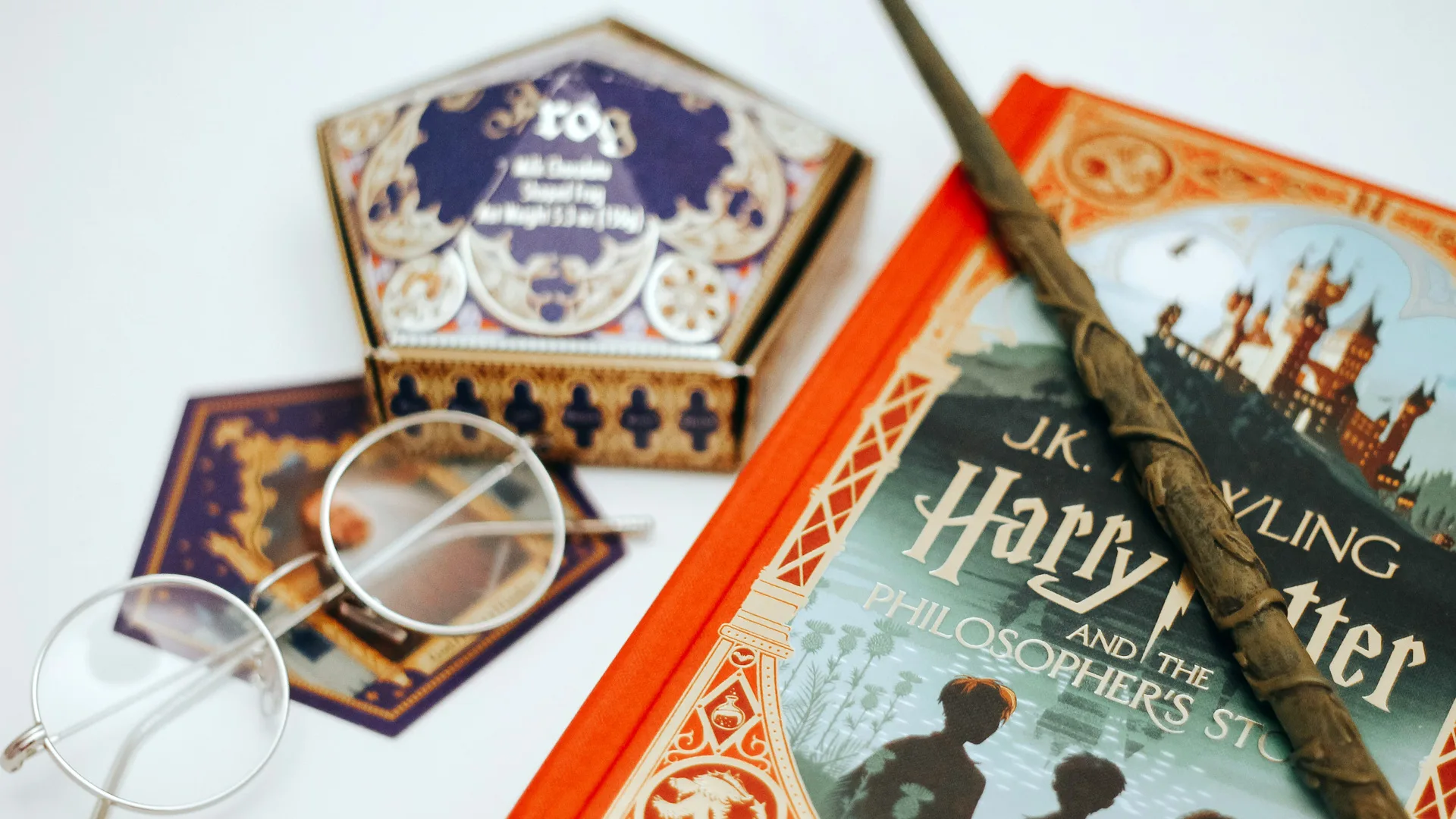 A photo of a number of Harry Potter related objects including a chocolate frog box in purple and gold, a wand, a pair of round glasses and a copy of The Philosopher's Stone book all on a white background.