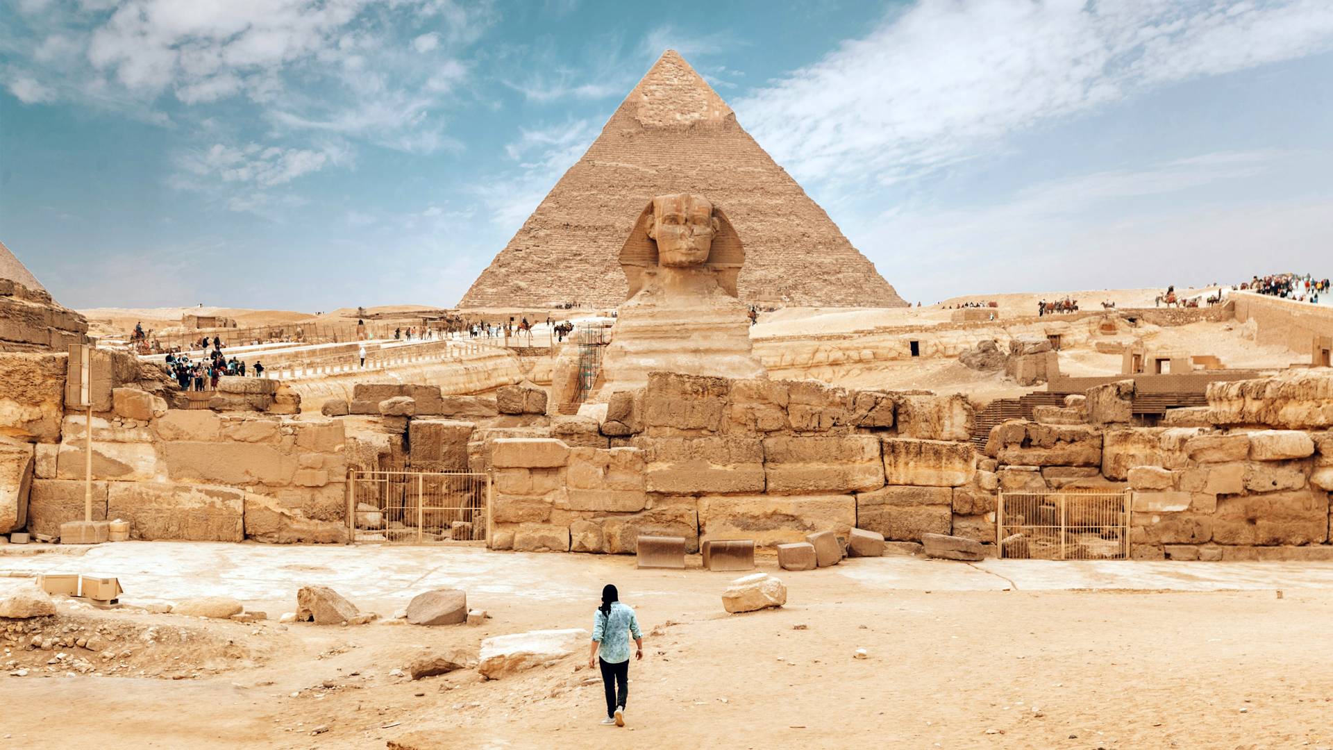 A photo of one of the pyramids in Egypt with a Sphynx sat outside, atop a pile of yellow bricks. In the foreground. a woman with a blue shirt and black trousers stands in the desert sand. The sky is blue with whispy white clouds.