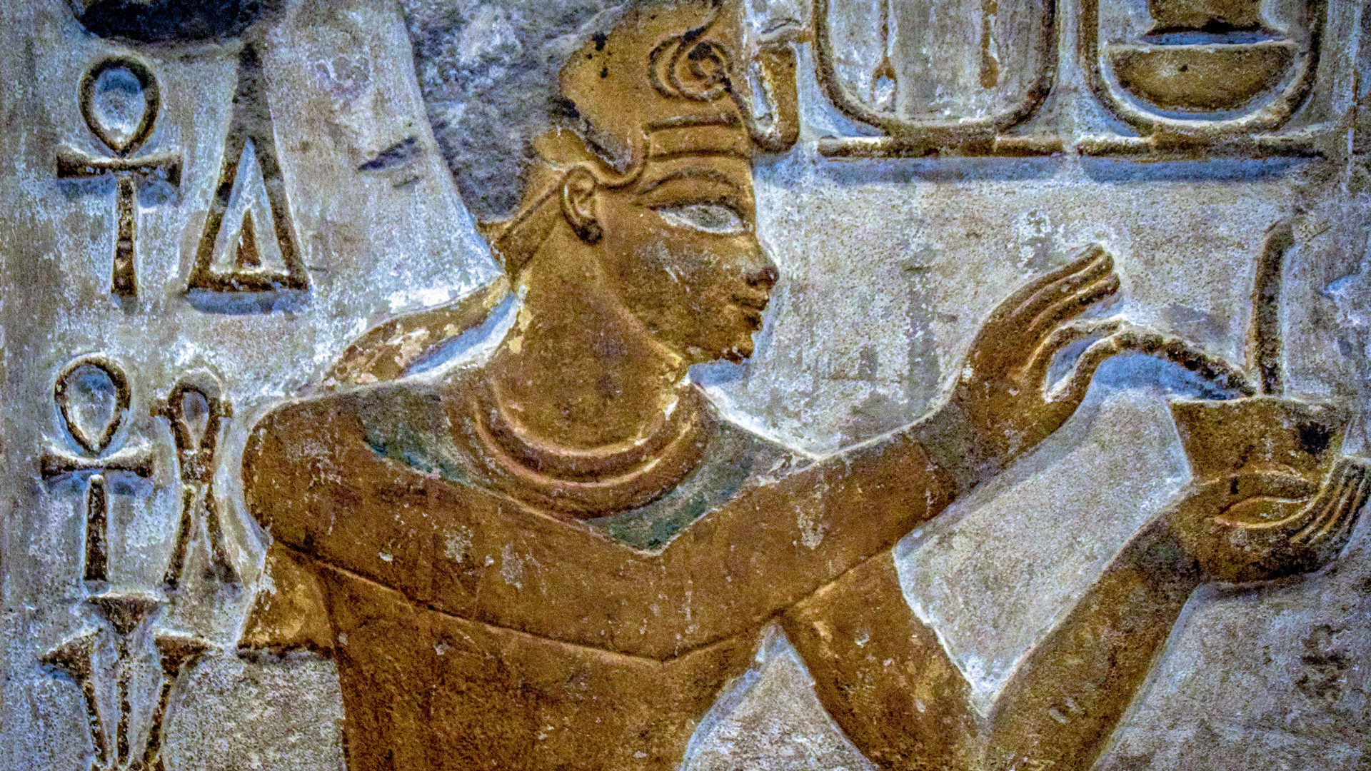 A photo of an Ancient Egyptian carving of a God in gold holding what looks like a pot of snakes with hieroglyphs surrounding him on a beige stone background.