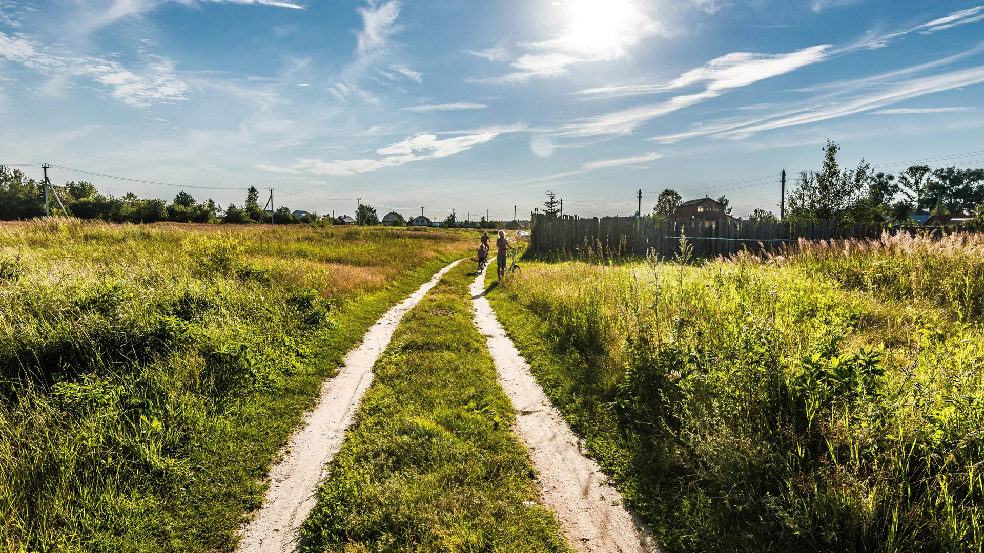 A photograph of the green countryside with a path leading off into the horizon. The sky is blue with the sun shining and two people are walking in the distance.