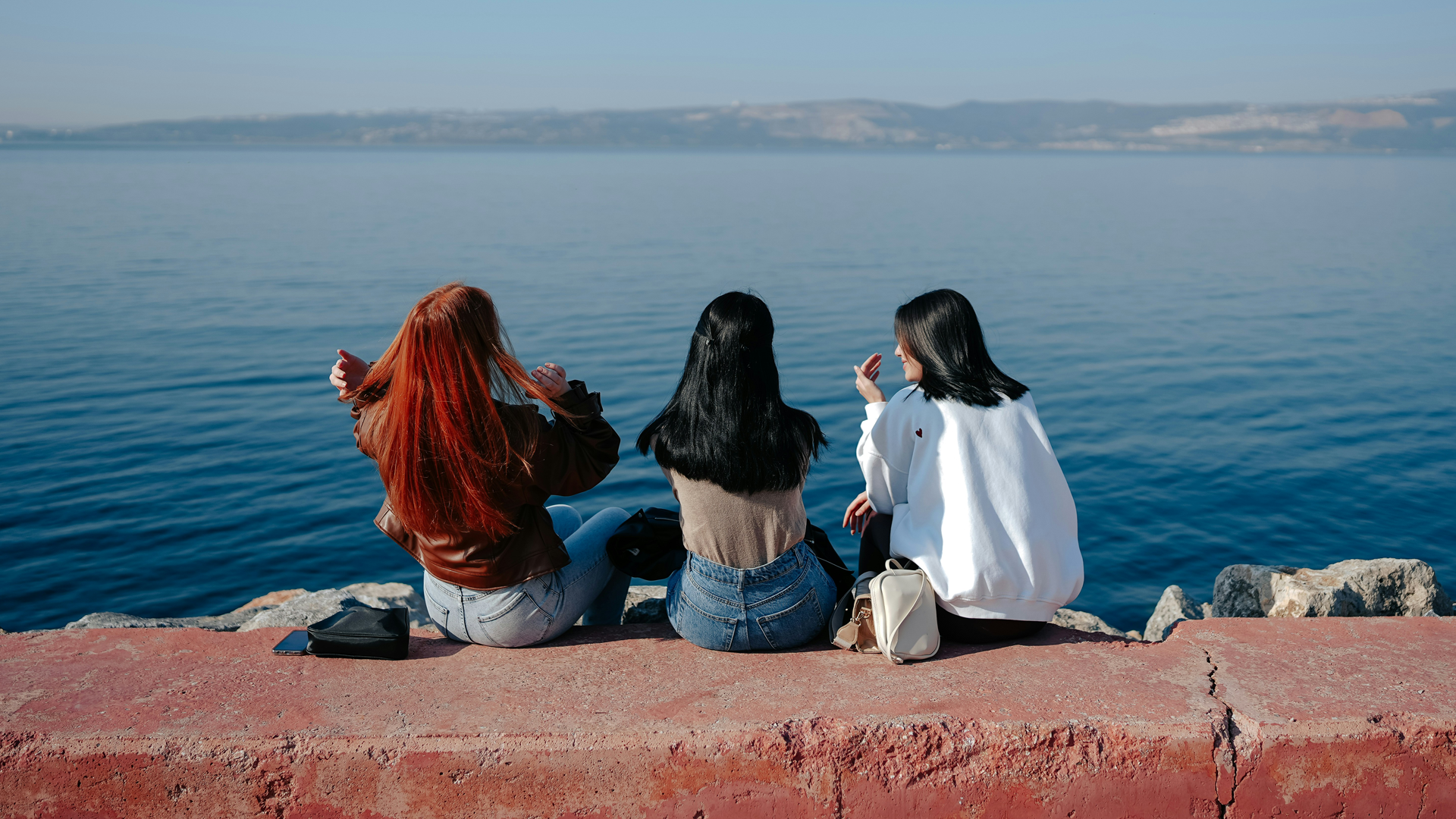 A group photo of three women sat on a red brick sea wall looking at the blue sea.