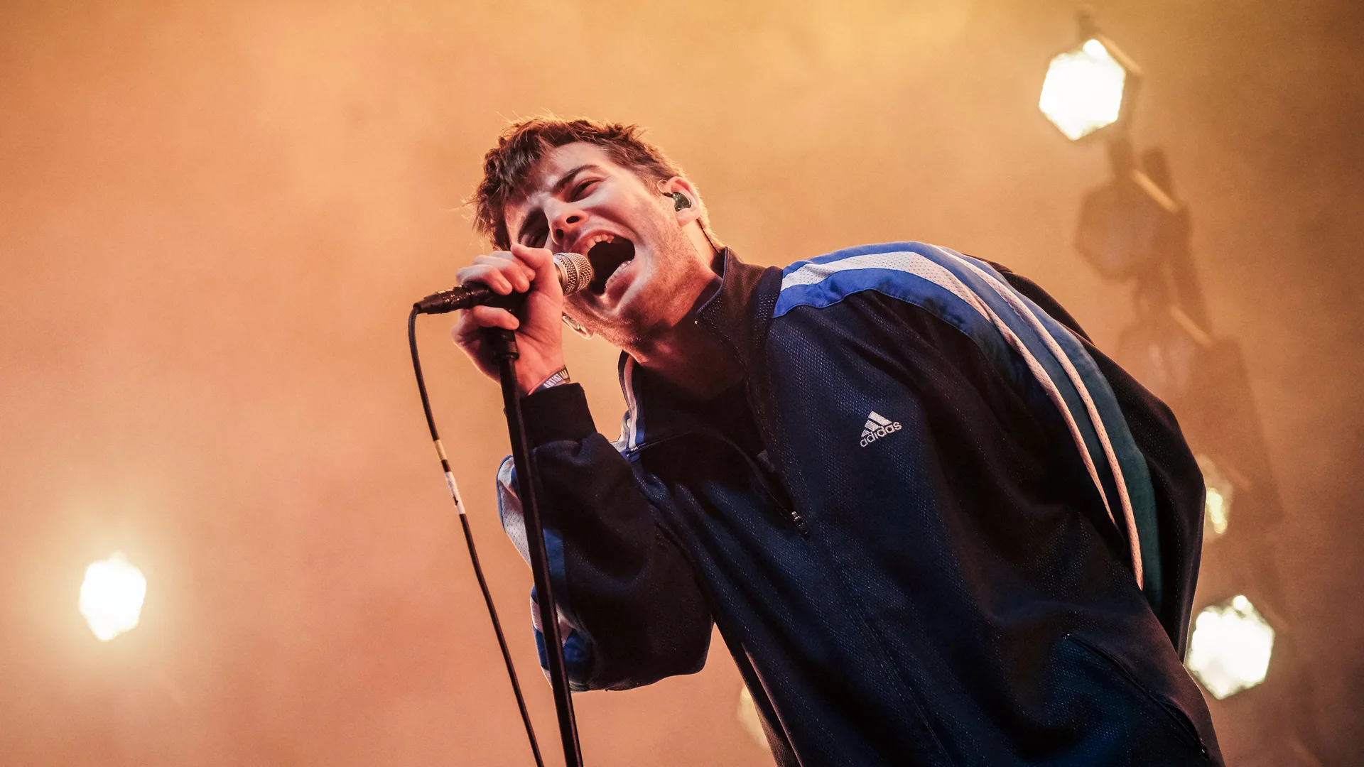 A photo of Fontaines DC singing at a festival on a stage with lights and orange mist. He is singing into the mic wearing a tracksuit top.