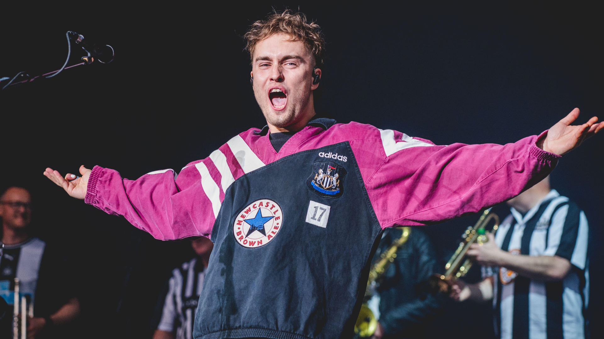 A photo of singer Sam Fender standing on stage with his arms stretched out wearing a Newcastle shirt in navy and pink with a shouting expression on his face against a black background.