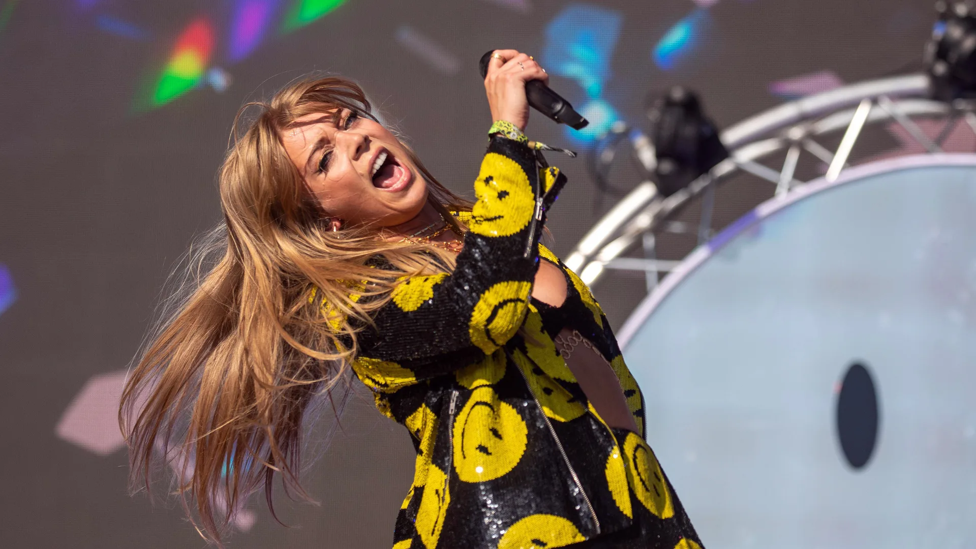 A photo of Becky Hill performing on stage throwing her head back holding onto the mic. She is wearing a black and yellow jacket against stage lights and scaffolding.