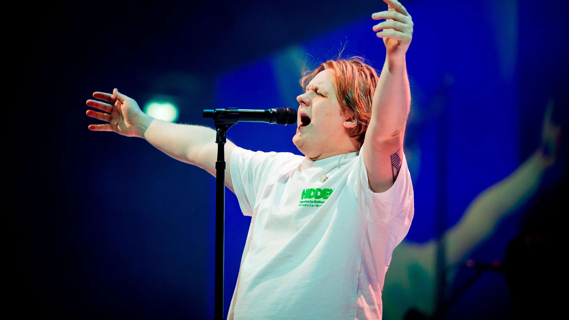 A photo of Lewis Capaldi performing on stage with his arms outstretched singing open-mouthed into the mic against a blue and black stage light.