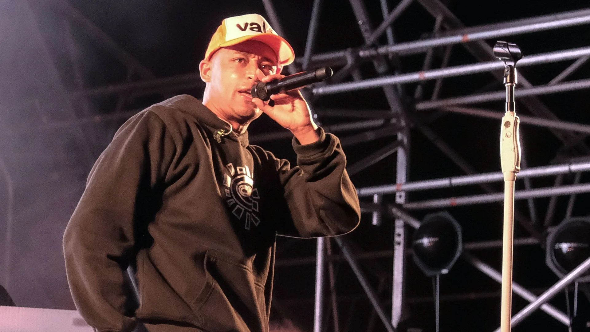 A photo of Loyle Carner performing on stage, holding the mic to his mouth. He is wearing a black hoodie and orange and white cap. Behind him is the stage with lighting scaffolding.