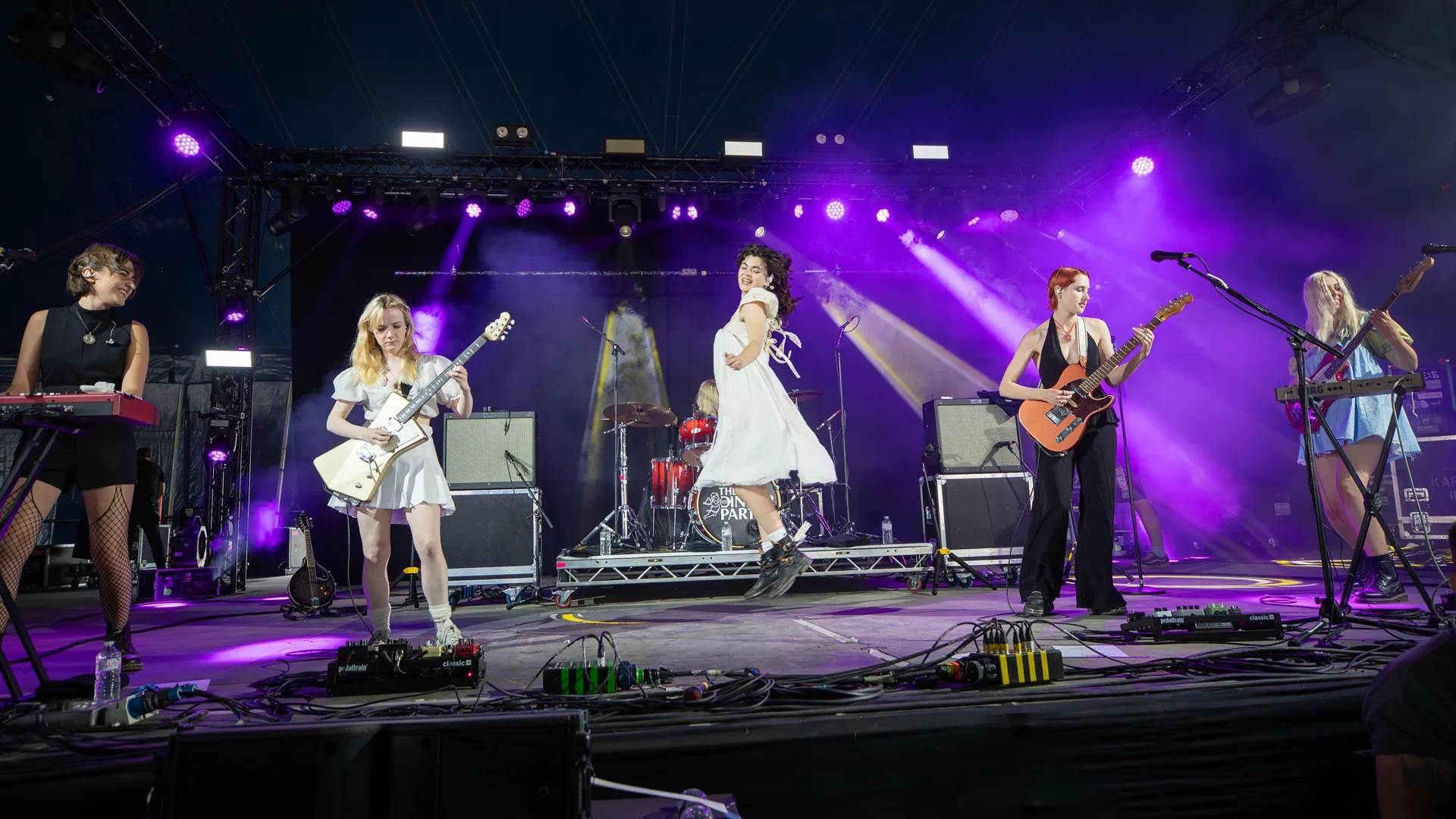 A photo of The Last Dinner Party performing on stage. All five members are showing left to right on stage with purple lights. The lead singer Abigail Morris is wearing a white dress.