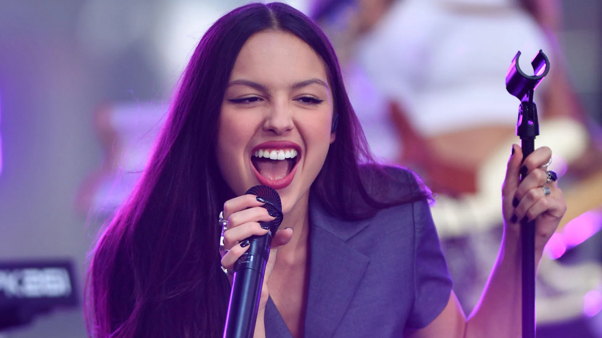 A photo of Olivia Rodrigo singing into a mic wearing a blue top against a purple lit background.