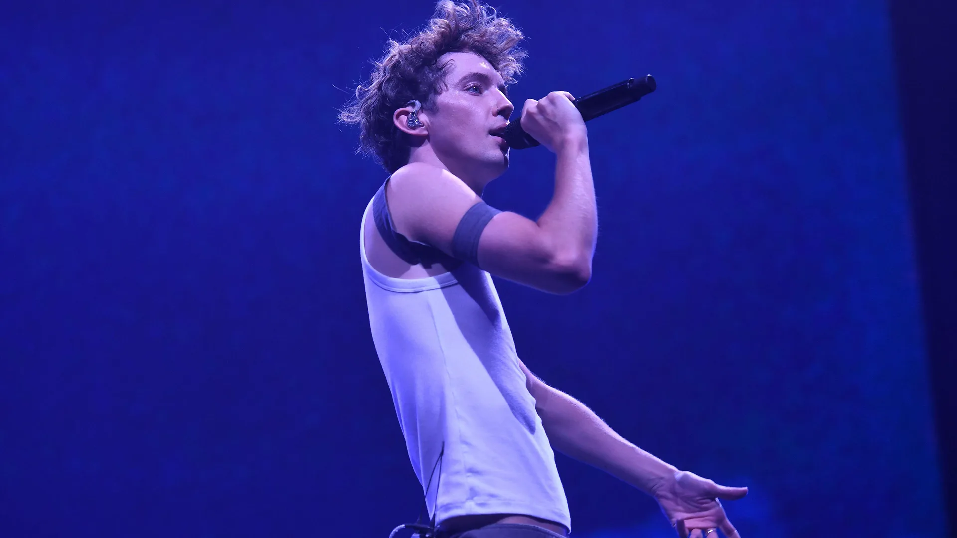 A photo of Troye Sivan performing on stage bathed in purple light. He is holding a mic to his mouth in a white vest top.