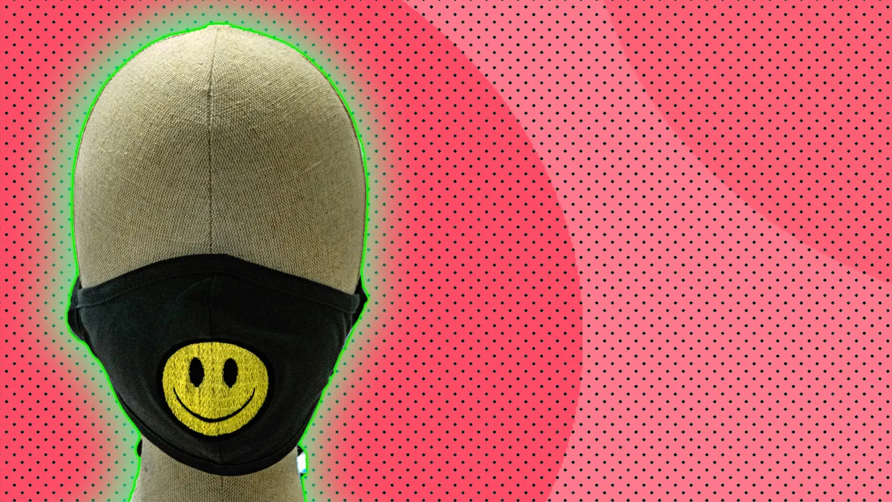 A photo of a mannequin head wearing a black mask with a yellow smiley face emoji on the front. It is against a pink dotted background.